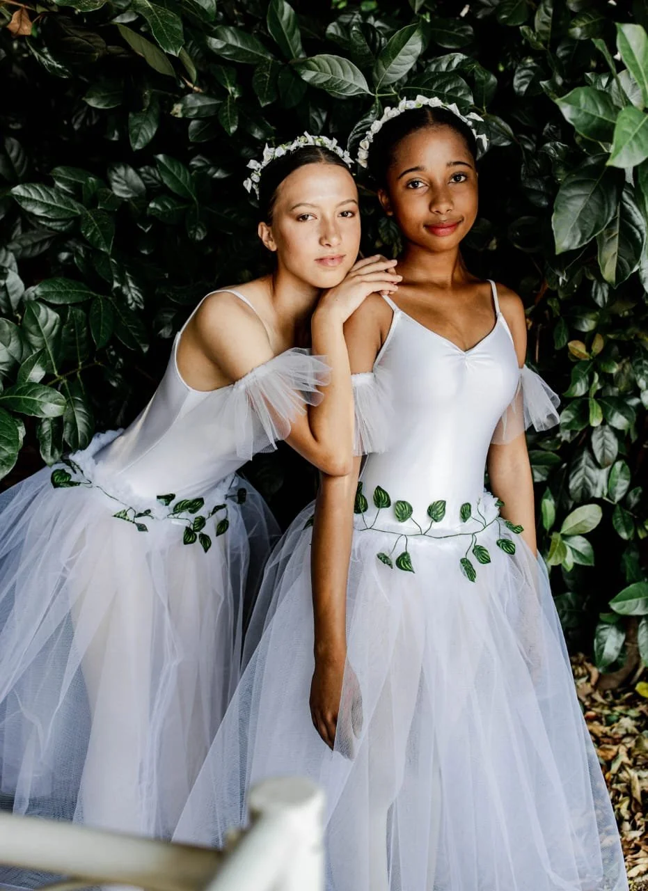 Two young women dressed as fairies in white dresses with leaf decorations, standing close together amidst lush green foliage, wearing floral headbands.