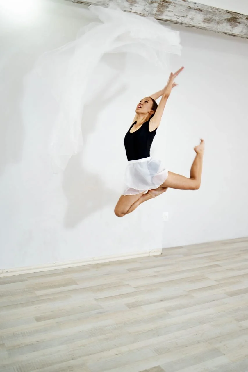 Ballet dancer mid-air leap in a room with white walls and light-colored wooden floor, wearing a black top and white tutu.