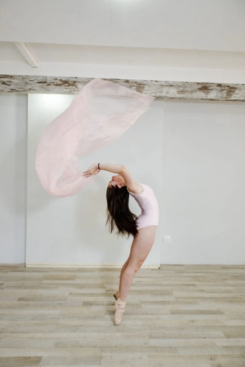 Ballet dancer in a pink leotard performing a pose with a flowing pink fabric in a studio with wooden flooring and white walls.