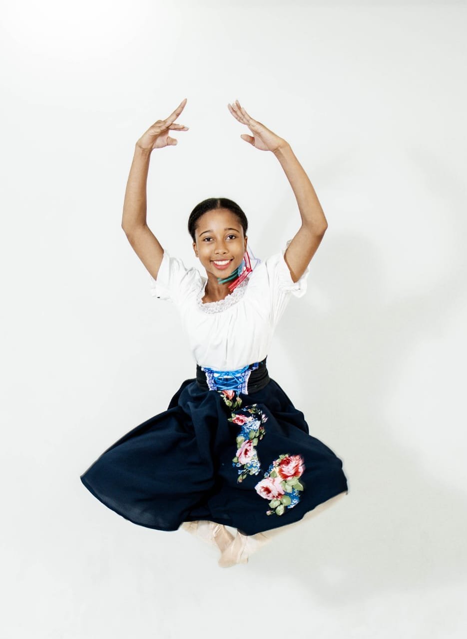 A young girl in traditional dress mid-air, smiling, with arms raised, against a plain white background.