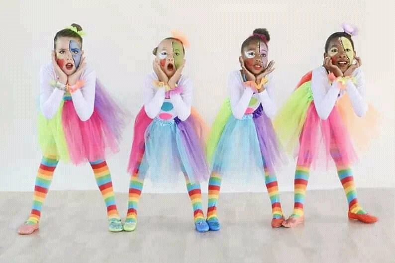 Four young girls dressed as clowns in colorful costumes with rainbow striped tights, tutus, and face paint, striking playful poses against a plain background.