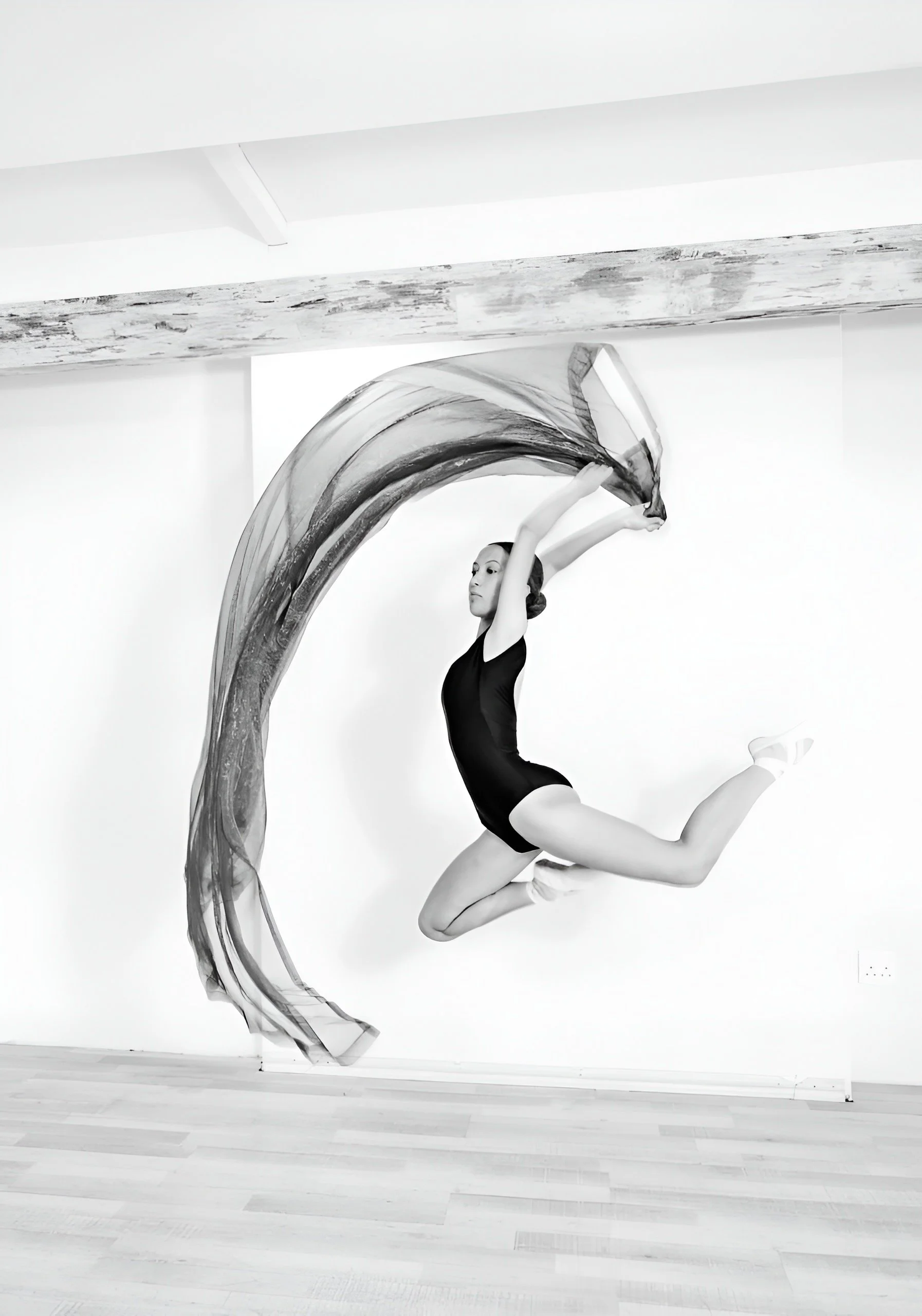 A ballerina performing a leap with an abstract scarf or fabric flowing around her, captured in a black and white minimalistic studio setting.