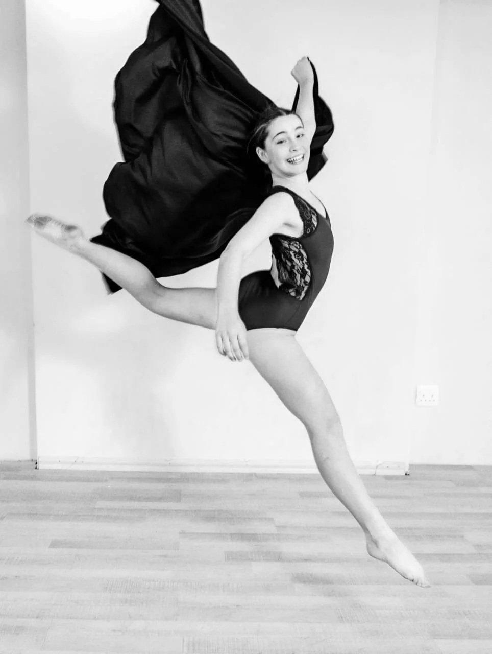 A young woman in a dance pose, mid-air with one leg extended behind her, one arm raised above her head, holding black fabric that flows behind her, wearing a black lace leotard, smiling, in a dance studio with a plain white wall.
