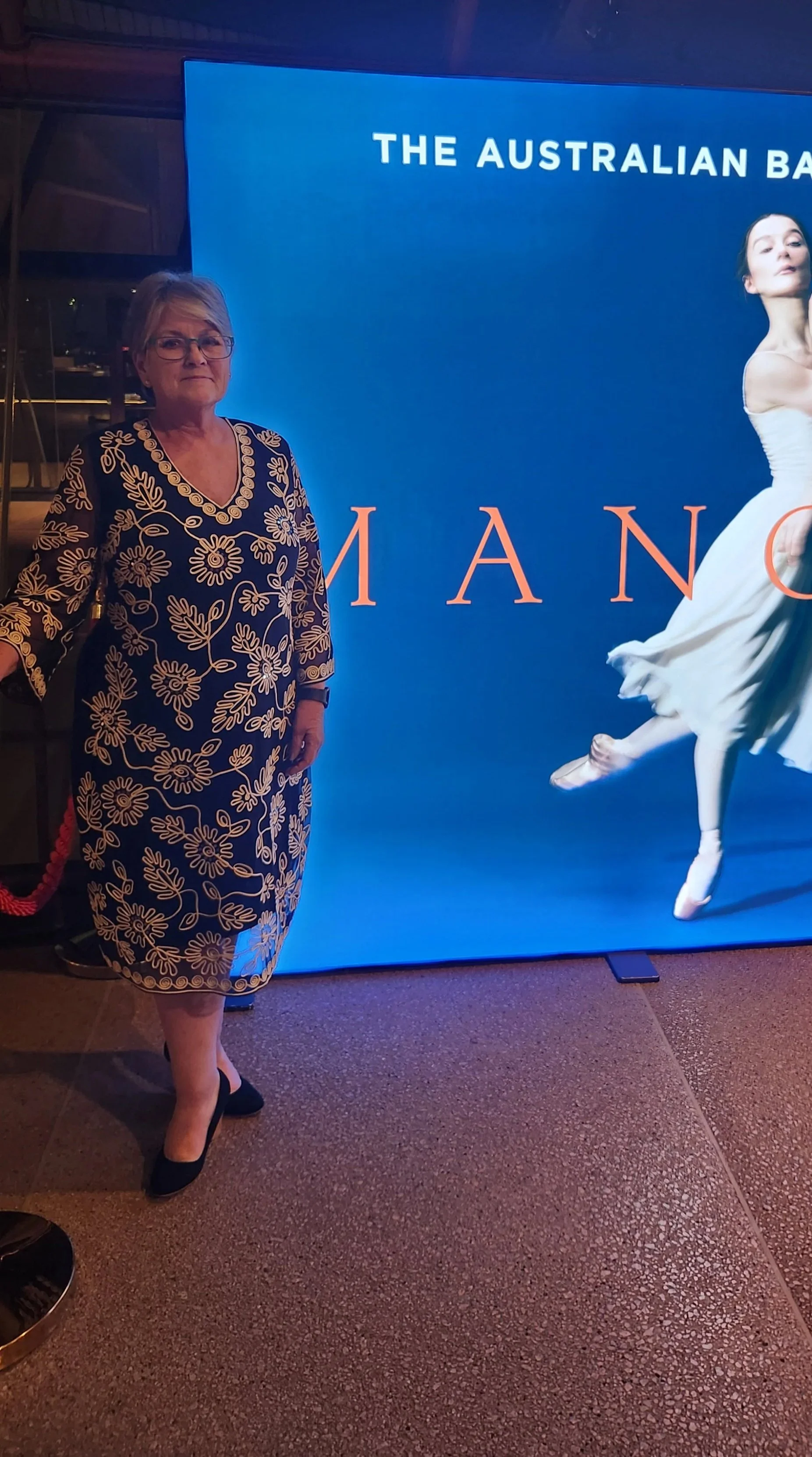 A woman standing next to a digital poster featuring a ballet dancer in a white dress, with part of the text 'THE AUSTRALIAN BA...'