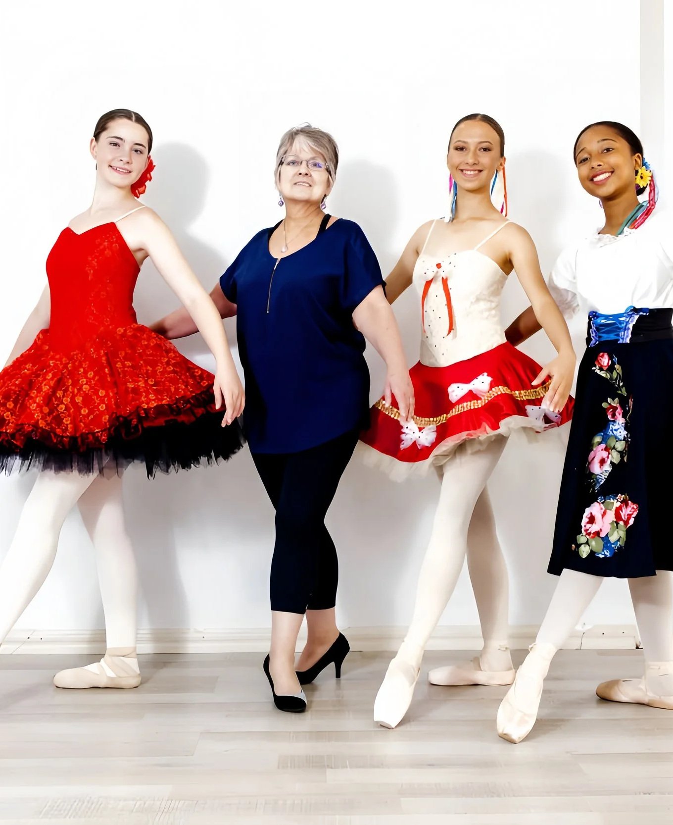 A woman standing among four ballet mannequins dressed in colorful costumes, all posed against a white wall.