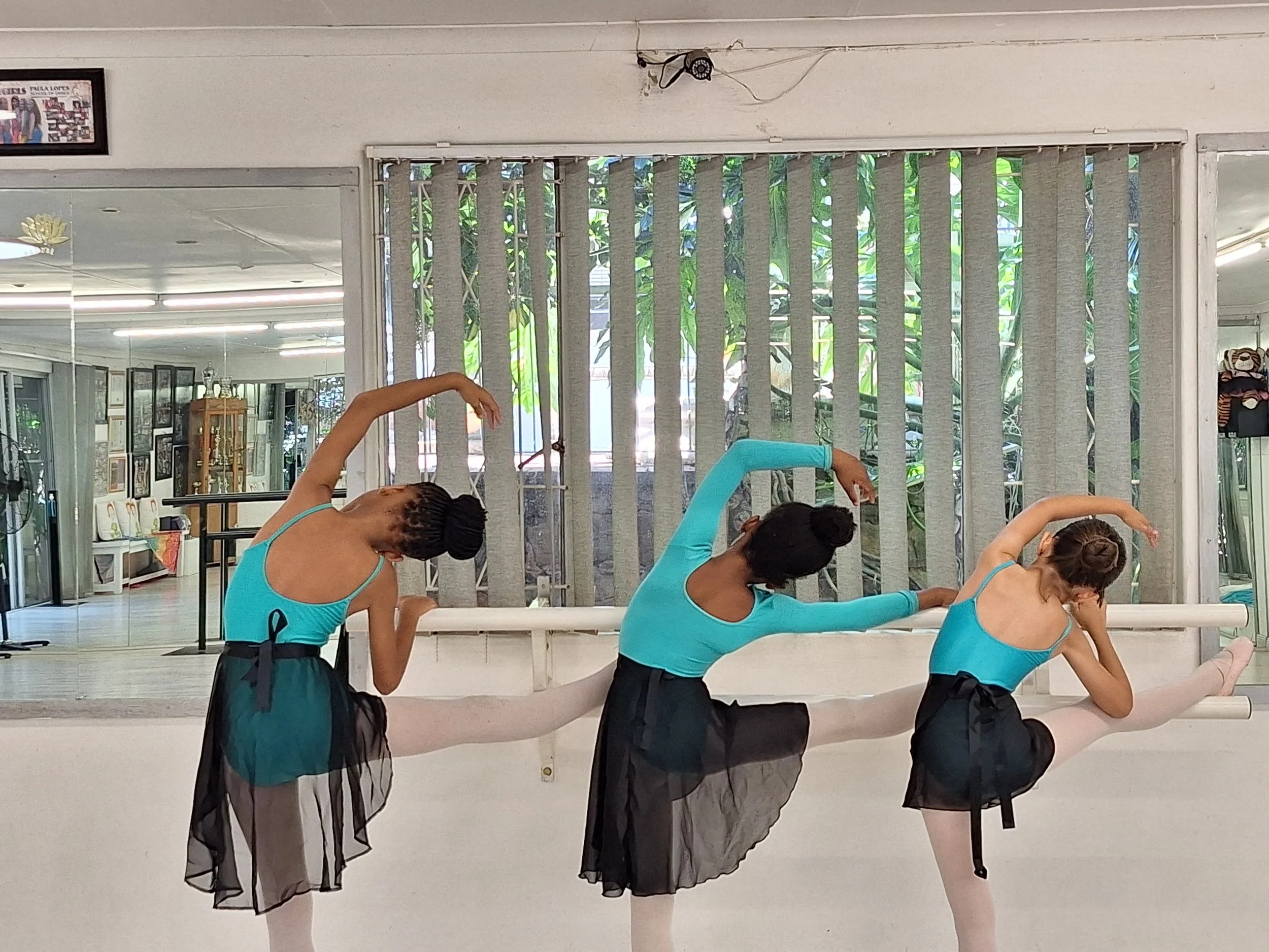 A group of ballet dancers practicing in a dance studio with large mirrors and vertical blinds, wearing blue leotards and black skirts.
