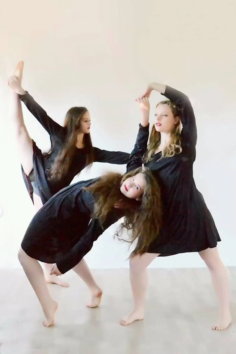 Three women in black dresses practicing ballet or dance poses indoors on a plain white wall background.