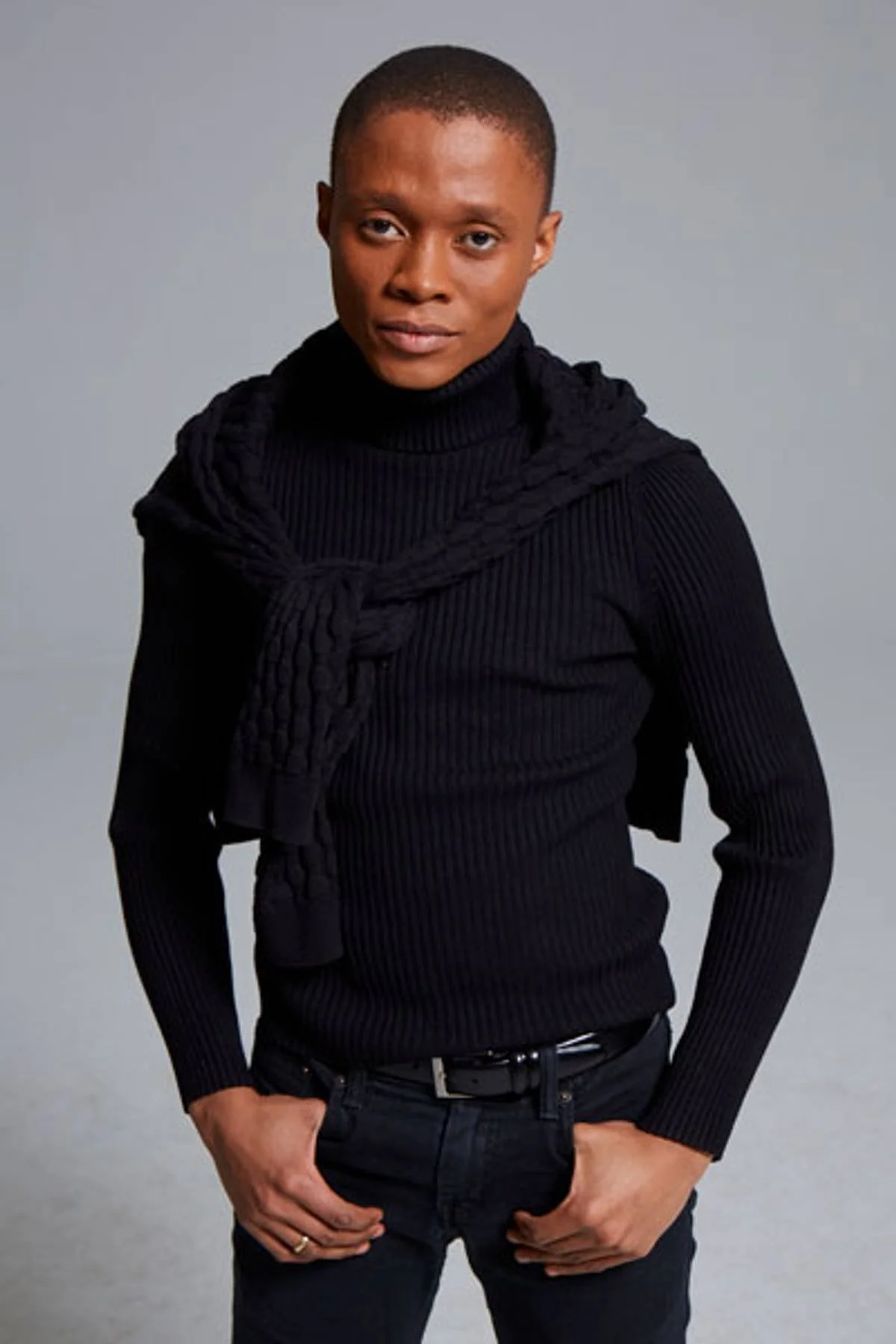 Person with short hairstyle wearing a black turtleneck sweater with a ribbed pattern, a black sweater tied around the shoulders, black jeans, and a black belt, standing against a plain gray background.