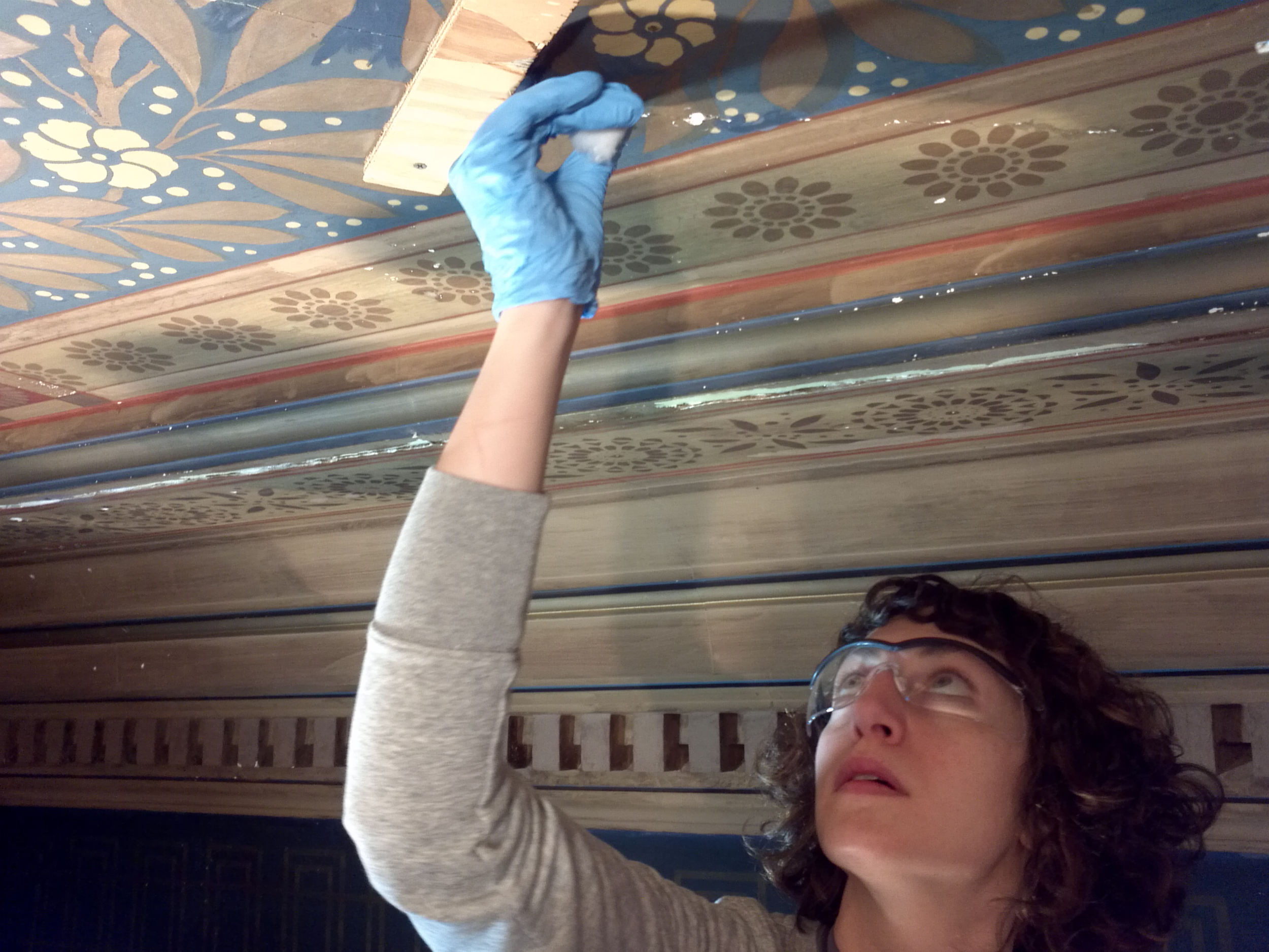 A woman with curly brown hair glasses and a gray long-sleeve shirt is reaching up with her right hand to tape a decorative wooden panel to a wall with floral patterns. The photo is upside down.