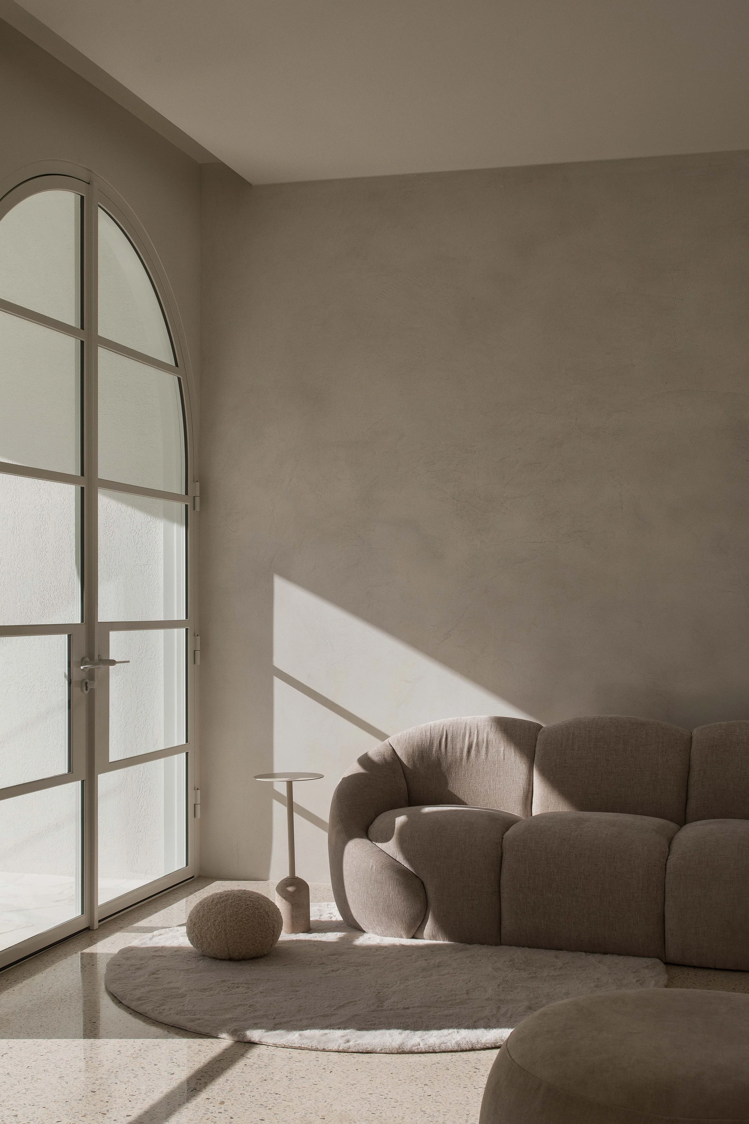 Living room with natural sunlight coming through arched window, a beige sofa, a round rug with a textured pouf, a standing side table, and minimalist decor in neutral tones.