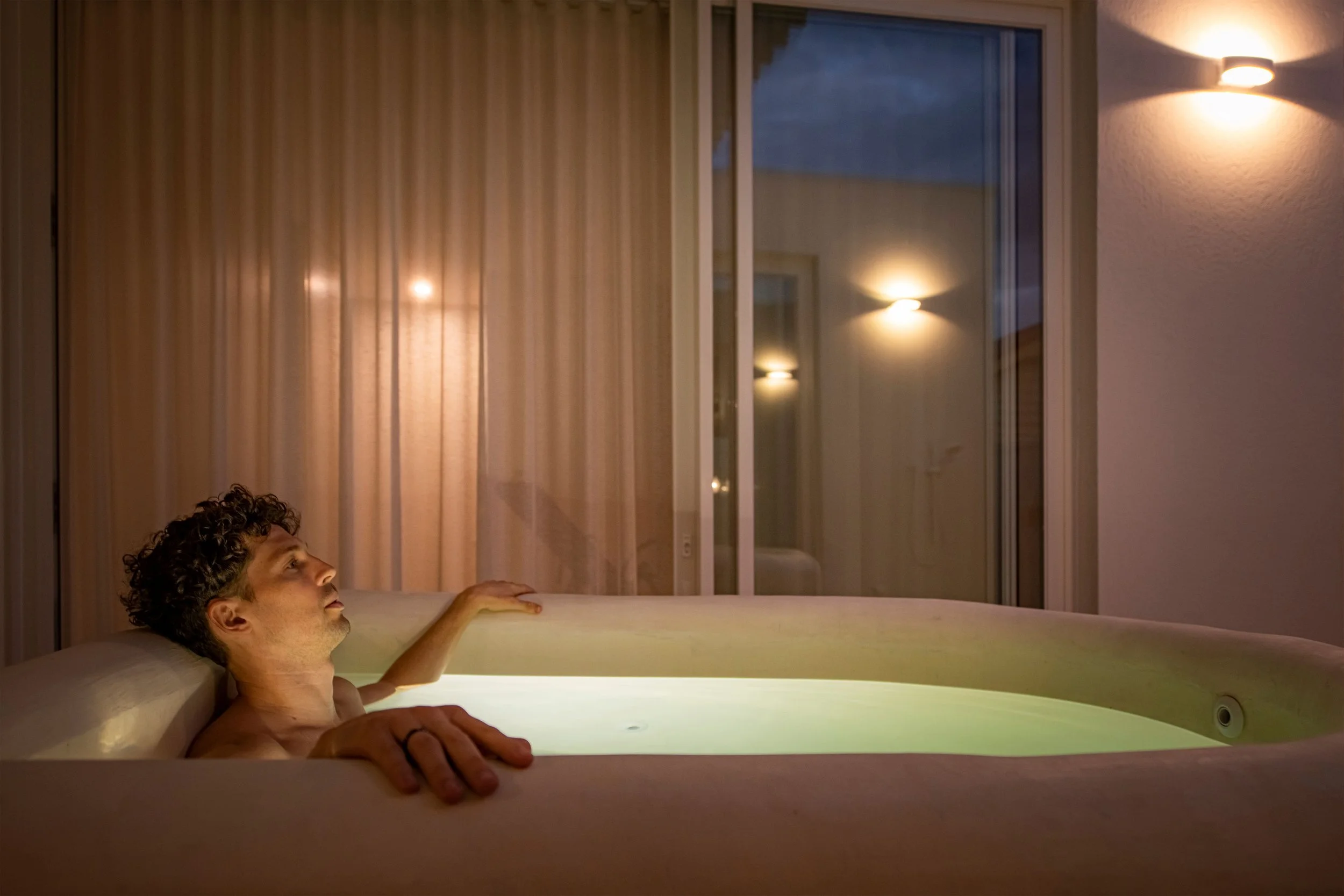 A man with curly hair relaxing in a modern bathtub with built-in lighting, surrounded by large windows with curtains, in a dimly lit room