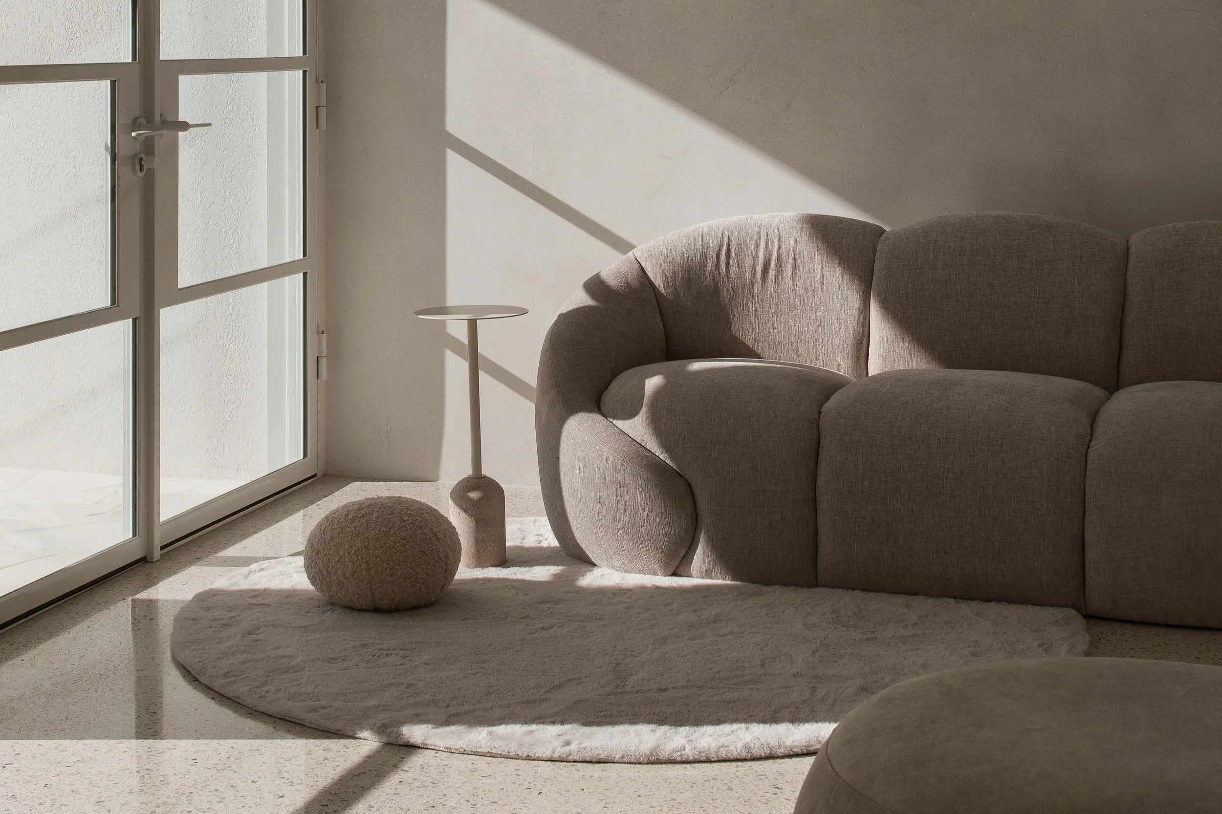 Sunlight streams through a glass door illuminating a beige sofa, a round beige ottoman, a small beige side table, a cream-colored rug, and a textured white wall in a minimalist living room.