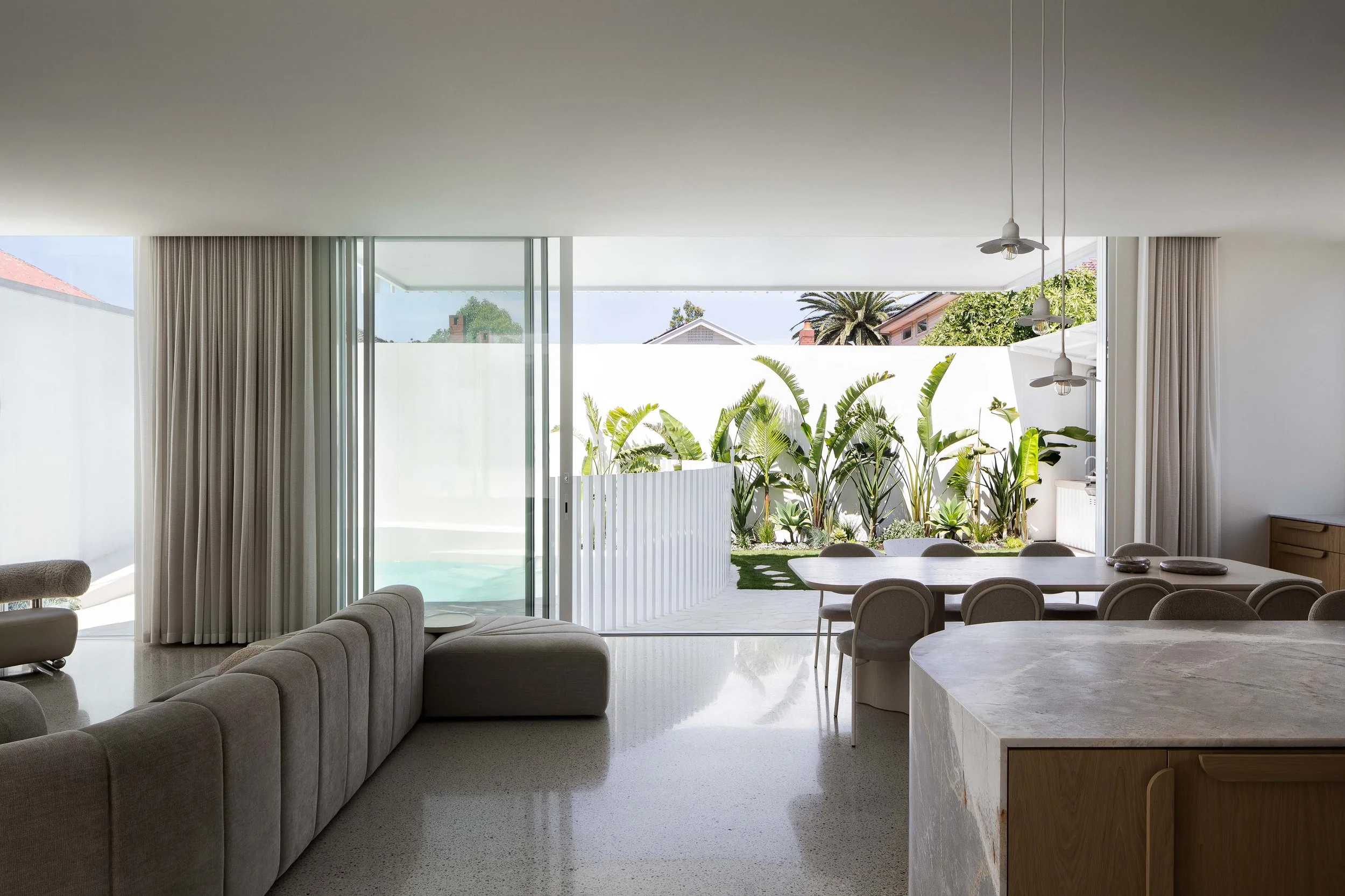 Contemporary living room with beige sectional sofa, marble kitchen island, and large sliding glass doors opening to a lush garden with tropical plants.