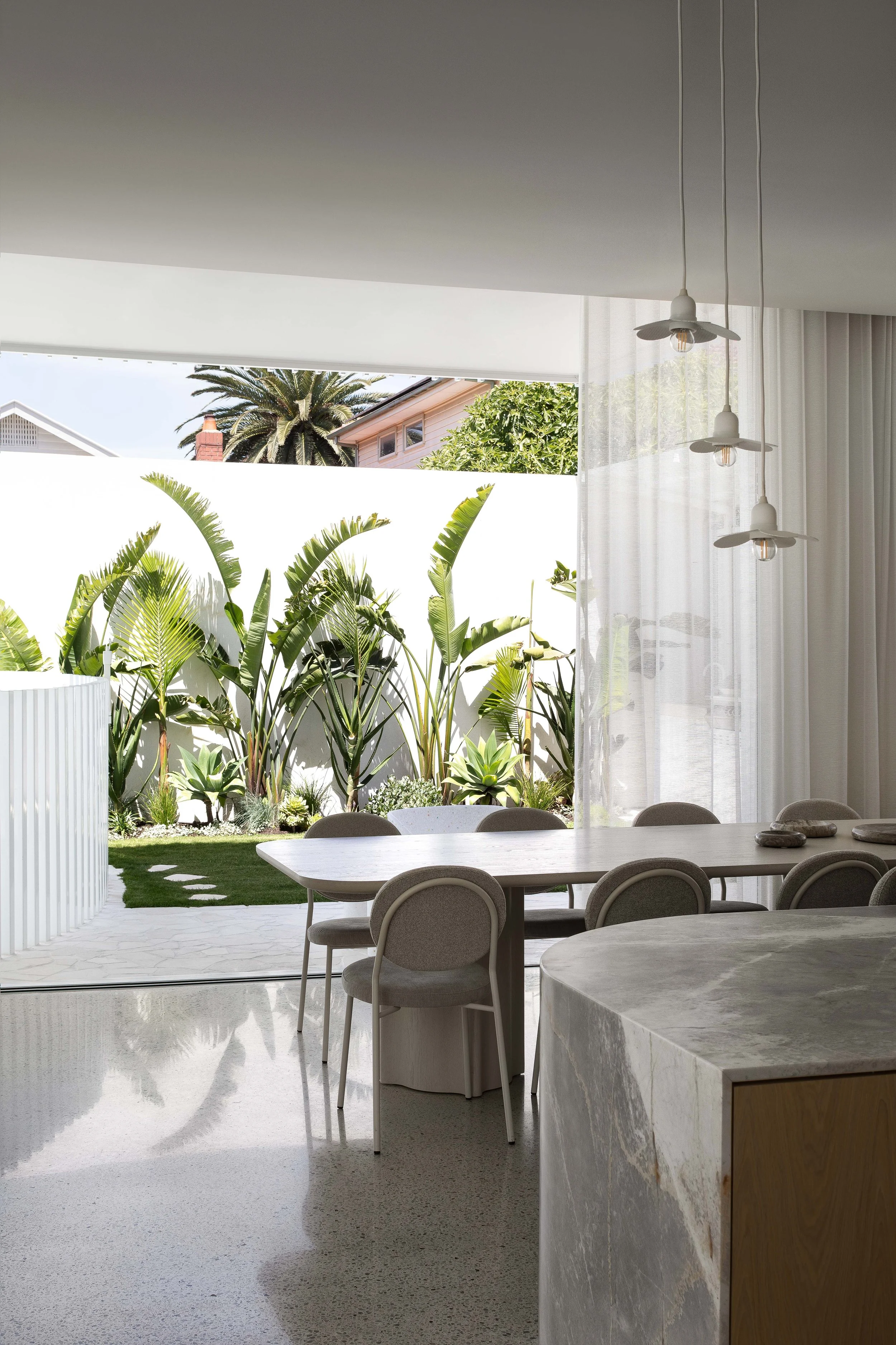Indoor dining area with marble table, chairs, and large glass sliding door leading to a garden with tropical plants and a view of neighboring houses with palm trees.