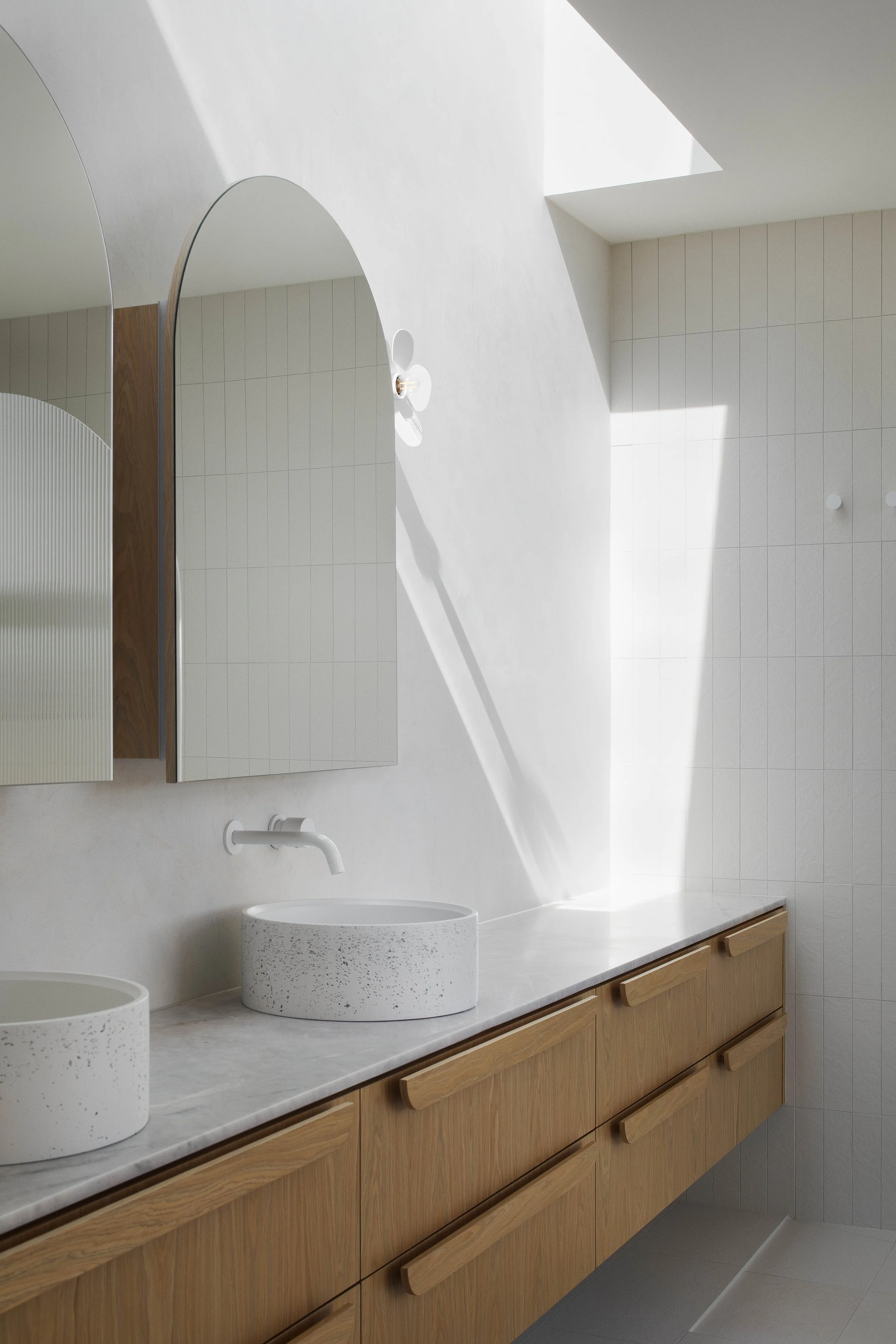 Modern bathroom with double oval sinks, wooden cabinetry, and a white wall mirror, illuminated by natural light from a skylight.