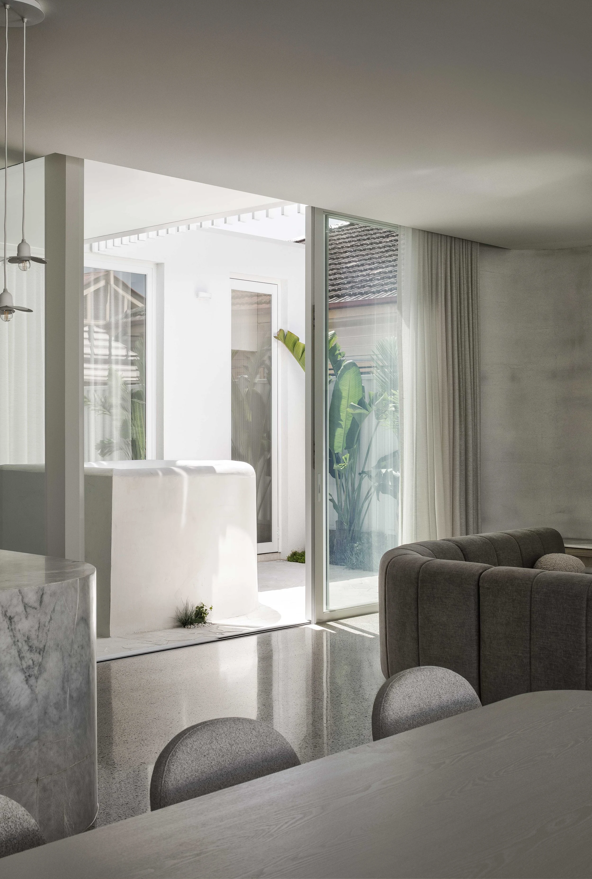Modern living room with sliding glass door leading to a patio and plants outside, partial view of a dining table and gray sofa, neutral color palette with natural light.