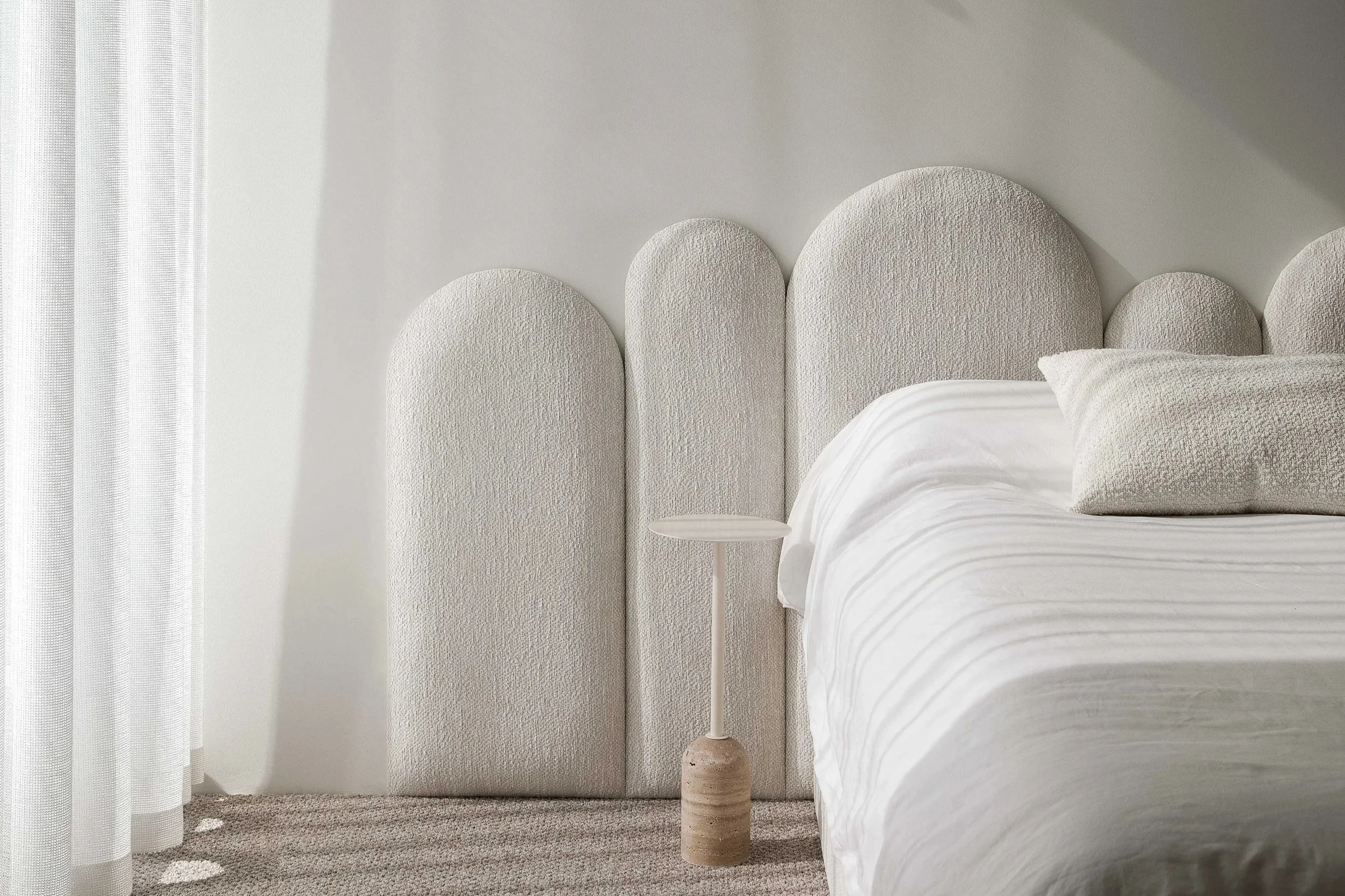 A bedroom with a white textured headboard, a white bed with pillows and a duvet, a small white side table, and sheer curtains on the left side.