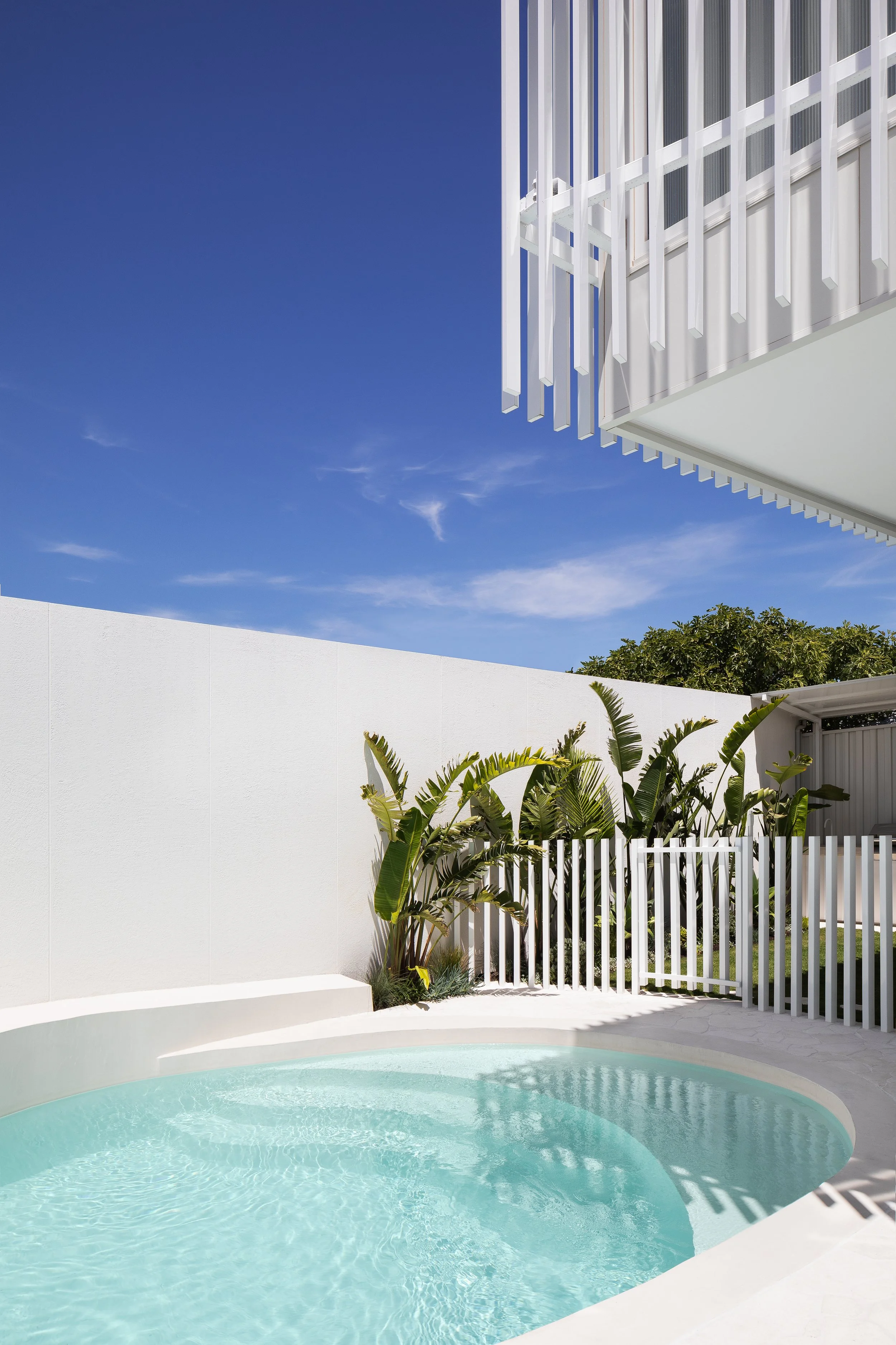 A modern house with a white fence, lush green plants, and a swimming pool with clear water under a blue sky.