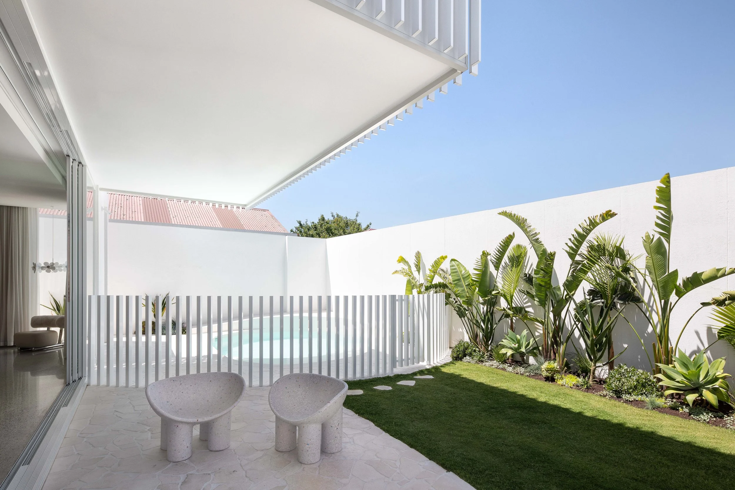Modern backyard patio with white concrete chairs, a white fence around a small pool or hot tub, lush green plants along the white wall, and a clear blue sky overhead.