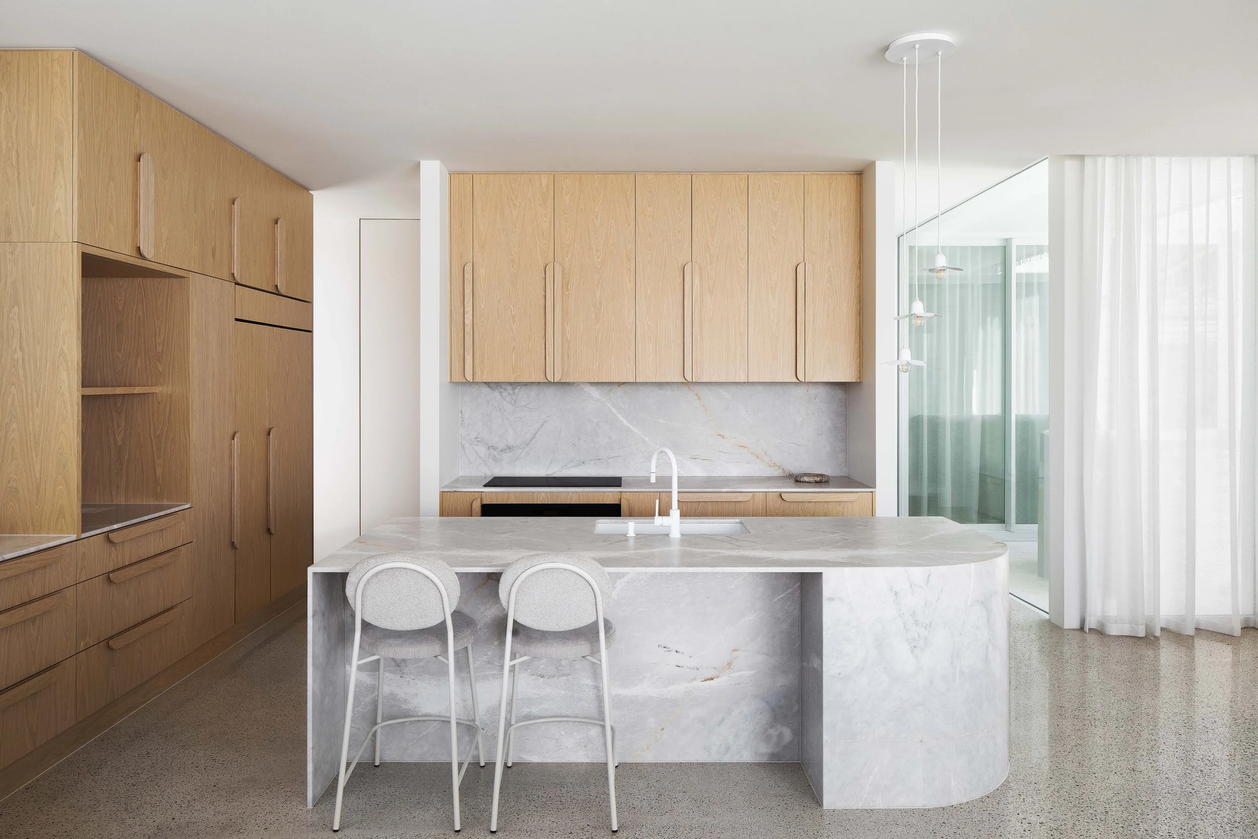 Modern kitchen with light wood cabinets, marble island, and white hanging pendant lights, adjacent to a glass sliding door with sheer white curtains.