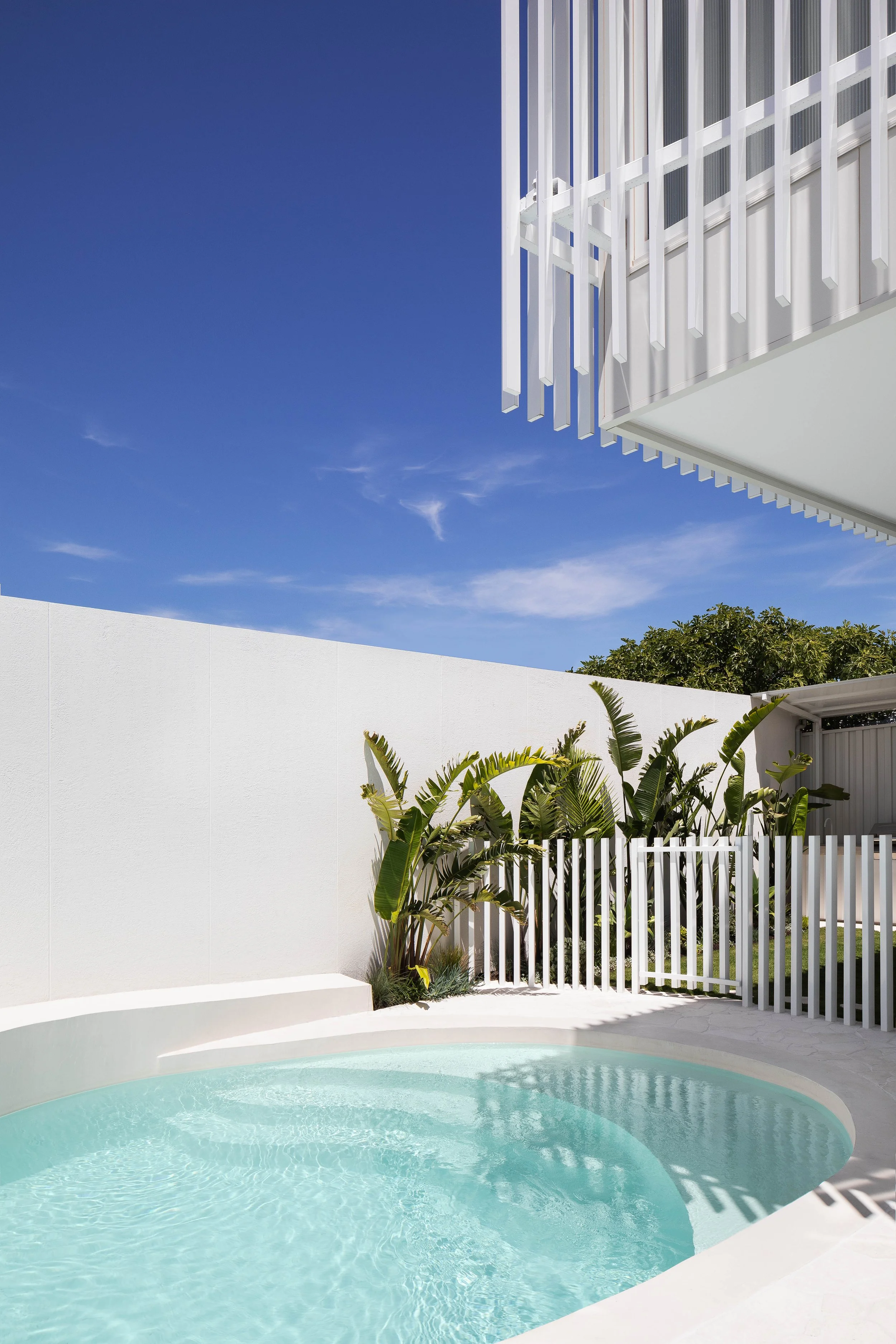 A modern backyard with a round swimming pool, white fence, green plants, and a white building with vertical slats, under a clear blue sky.