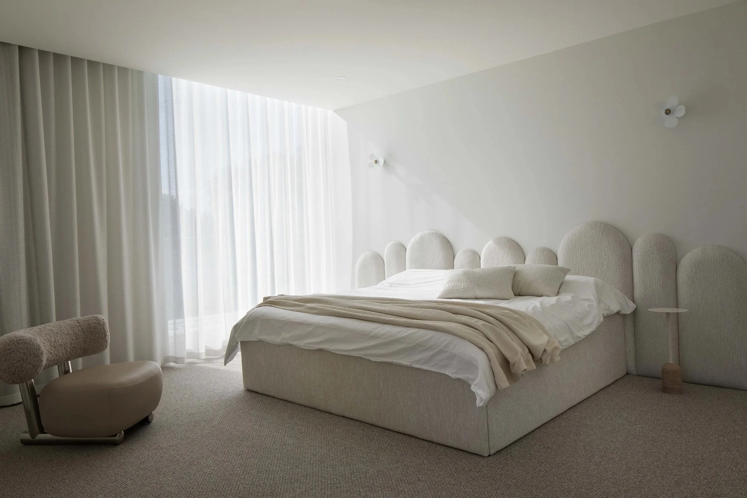 A modern bedroom with a white bed, round headboard, beige blanket, and white pillows. Next to the bed, there is a beige cushioned chair or stool. The room has large windows with sheer white curtains allowing natural light to fill the space.
