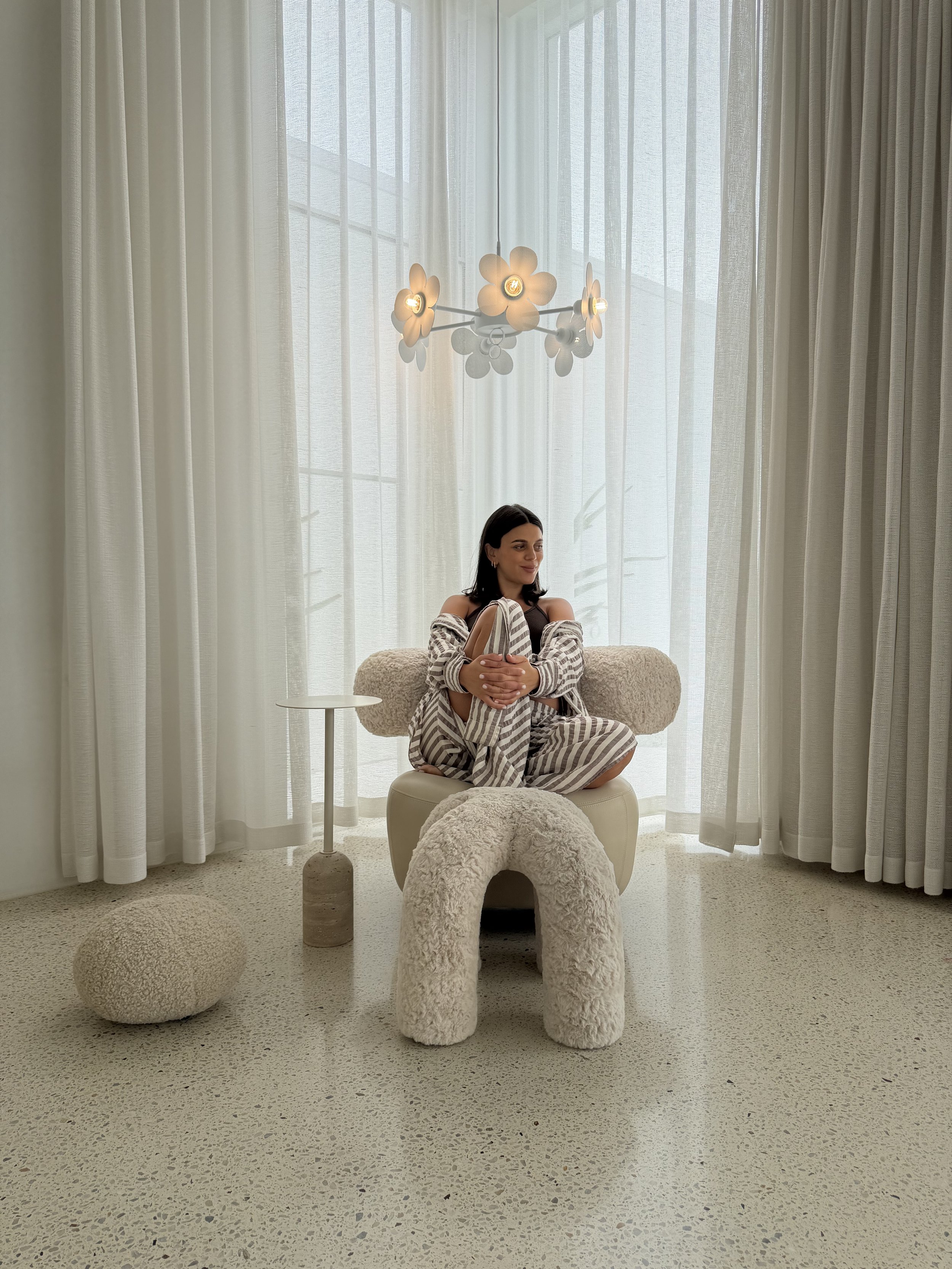 Interior scene with a woman sitting cross-legged on a fluffy armchair in front of large sheer curtains, with a modern floral chandelier above and textured flooring.