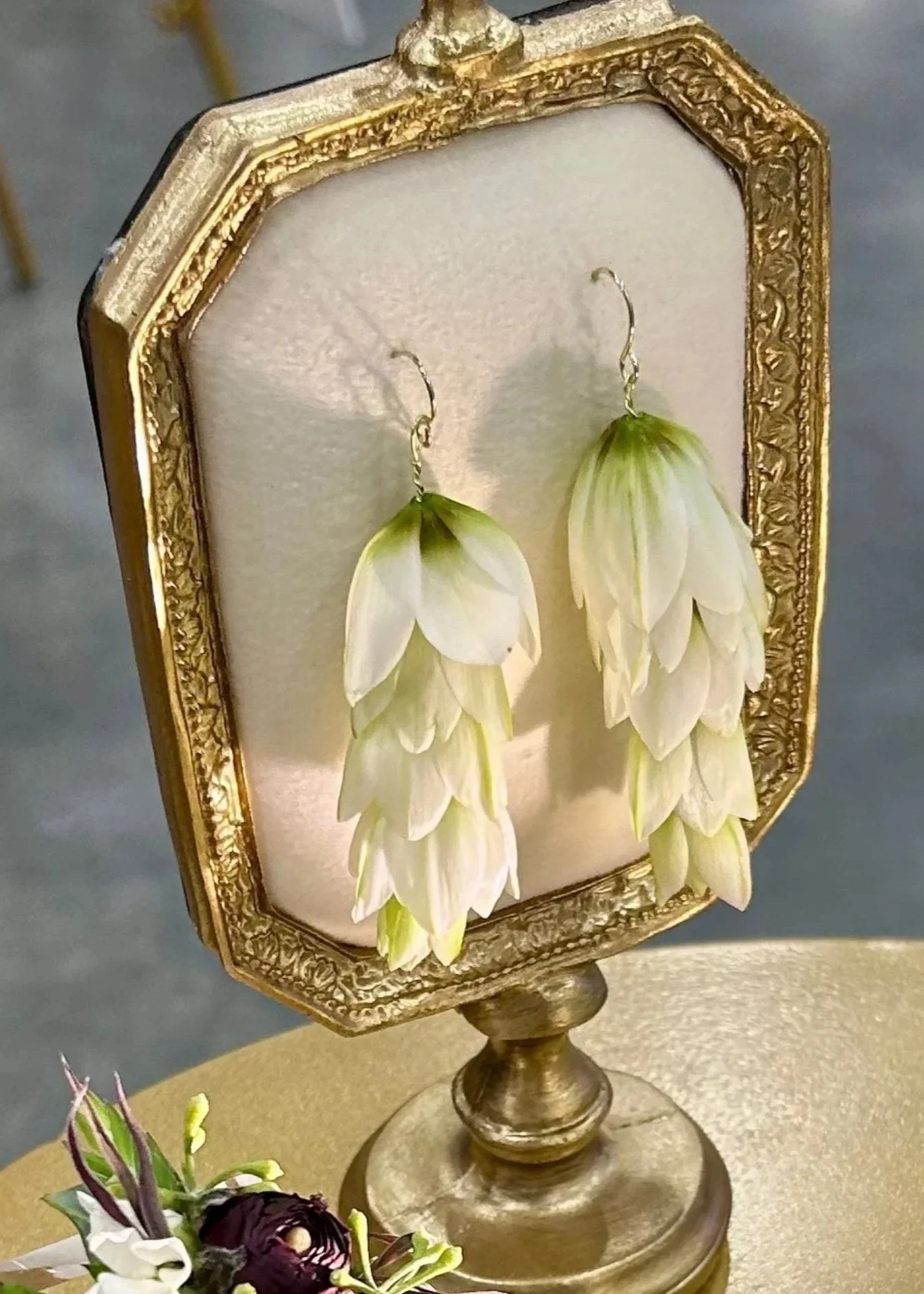 A pair of white, bell-shaped earrings made from real living flowers displayed on an ornate gold picture frame with a golden stand.