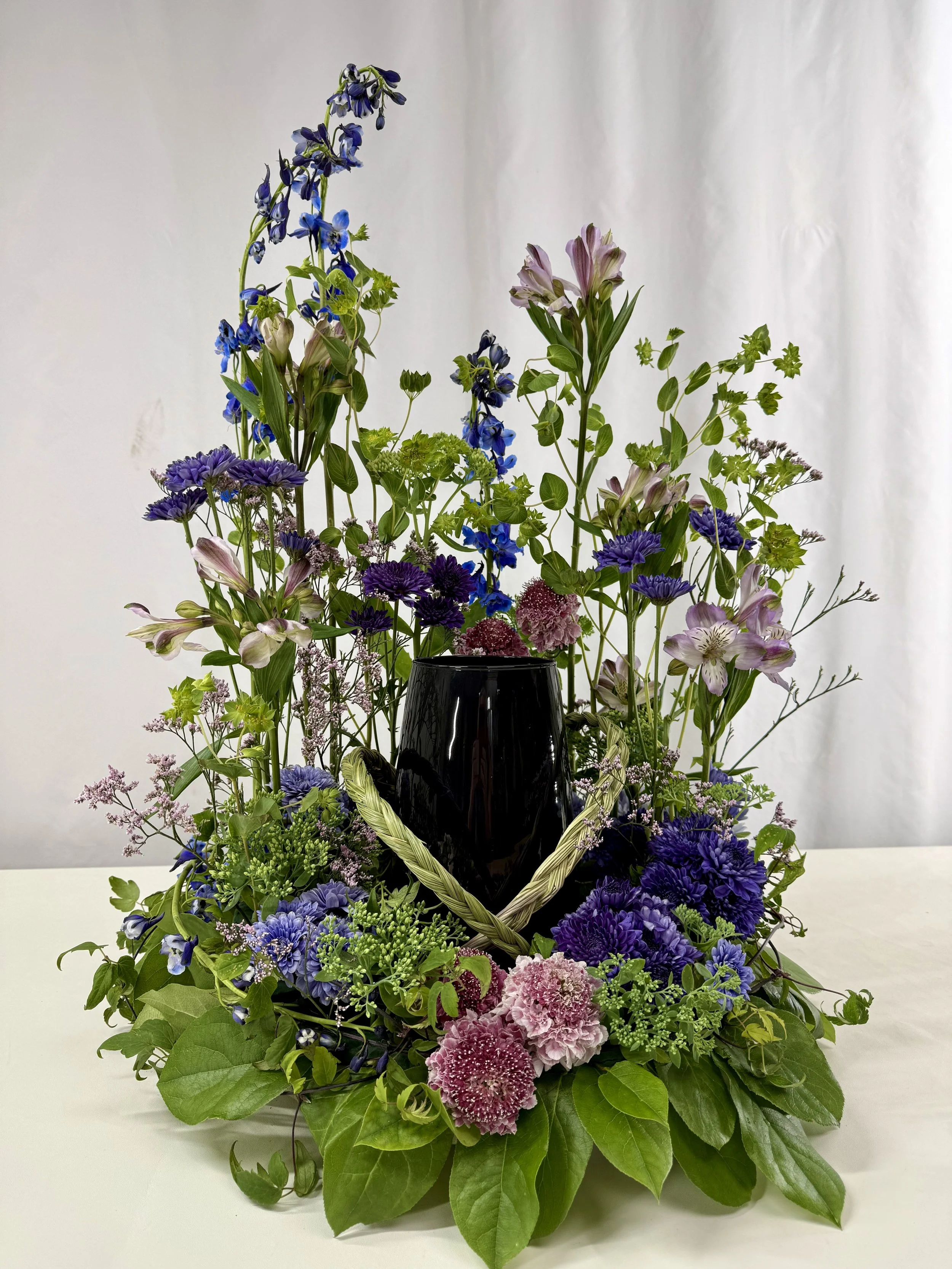 A sympathy meadow floral arrangement made for surrounding an urn with purple, pink, and white flowers, including delphiniums, mums and accented with greenery and braided sweetgrass around the urn's neck.