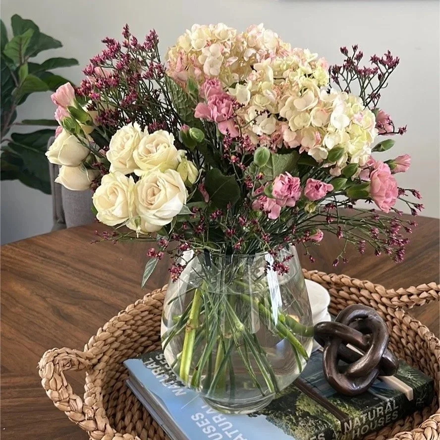 Cream roses, hydrangea and pink wax flower in a glass vase on a woven tray.