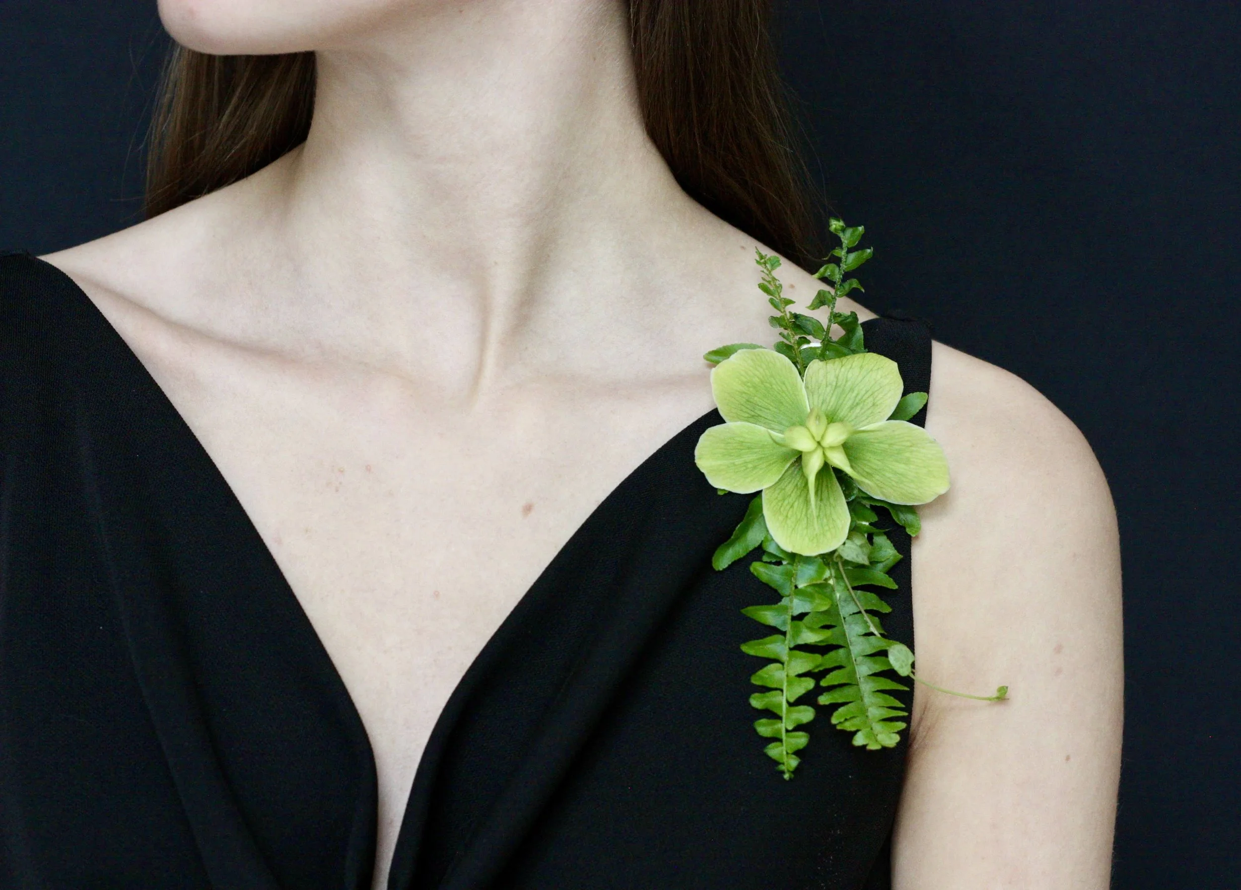 A woman wearing a black dress has a green hellebore flower and fern foliage pinned to her left shoulder.