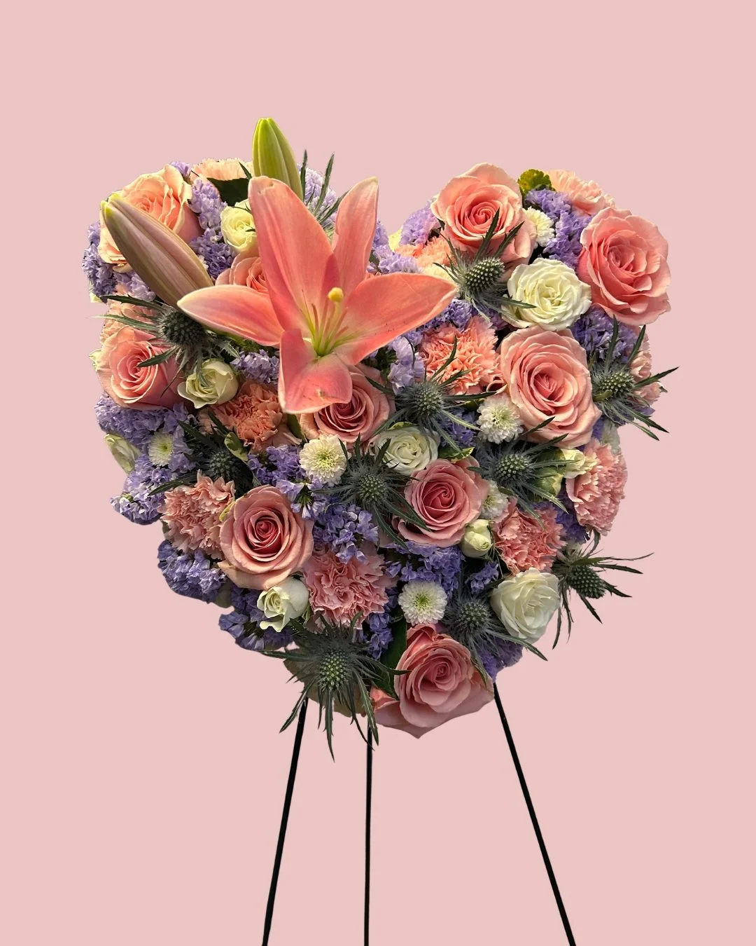 A floral heart-shaped sympathy arrangement with pink lilies, roses, purple statice, white spray roses and mums, and blue thistles on a pink background.