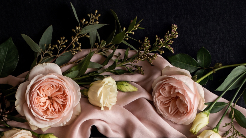 A floral arrangement with pink and white roses, green leaves, small buds, and greenery on a soft pink satin fabric.