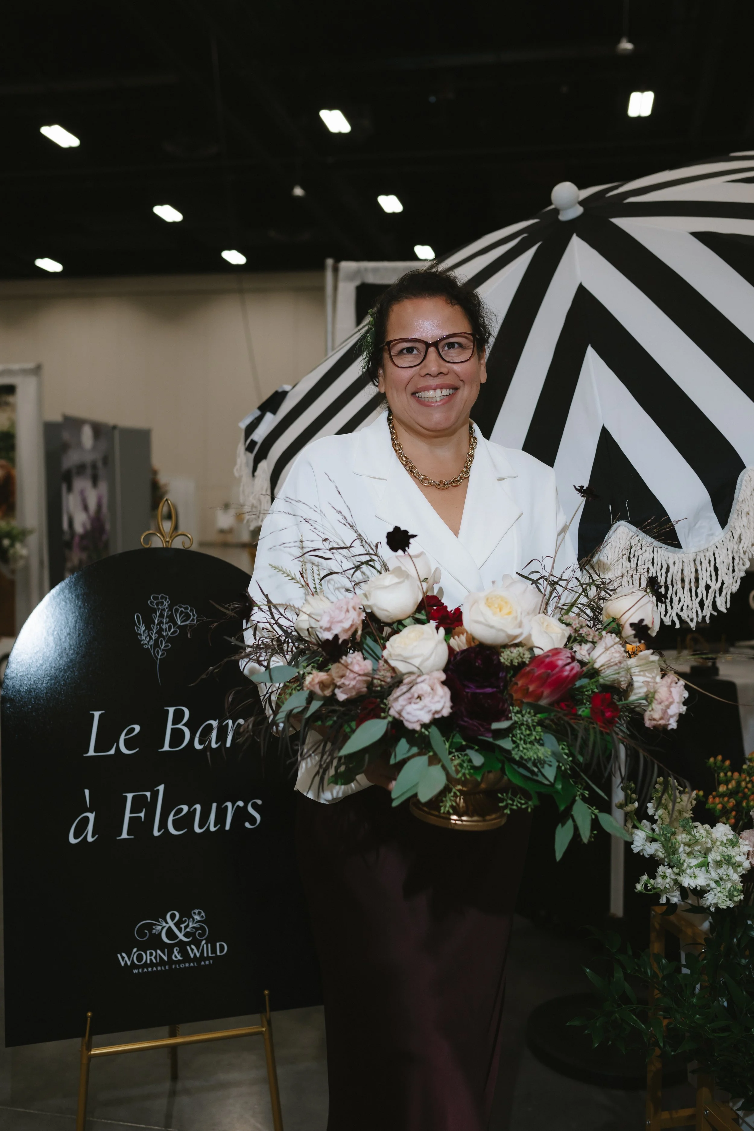 A woman in glasses and a white blazer smiling at a floral event, standing behind a large bouquet of flowers with a black and white striped umbrella in the background.