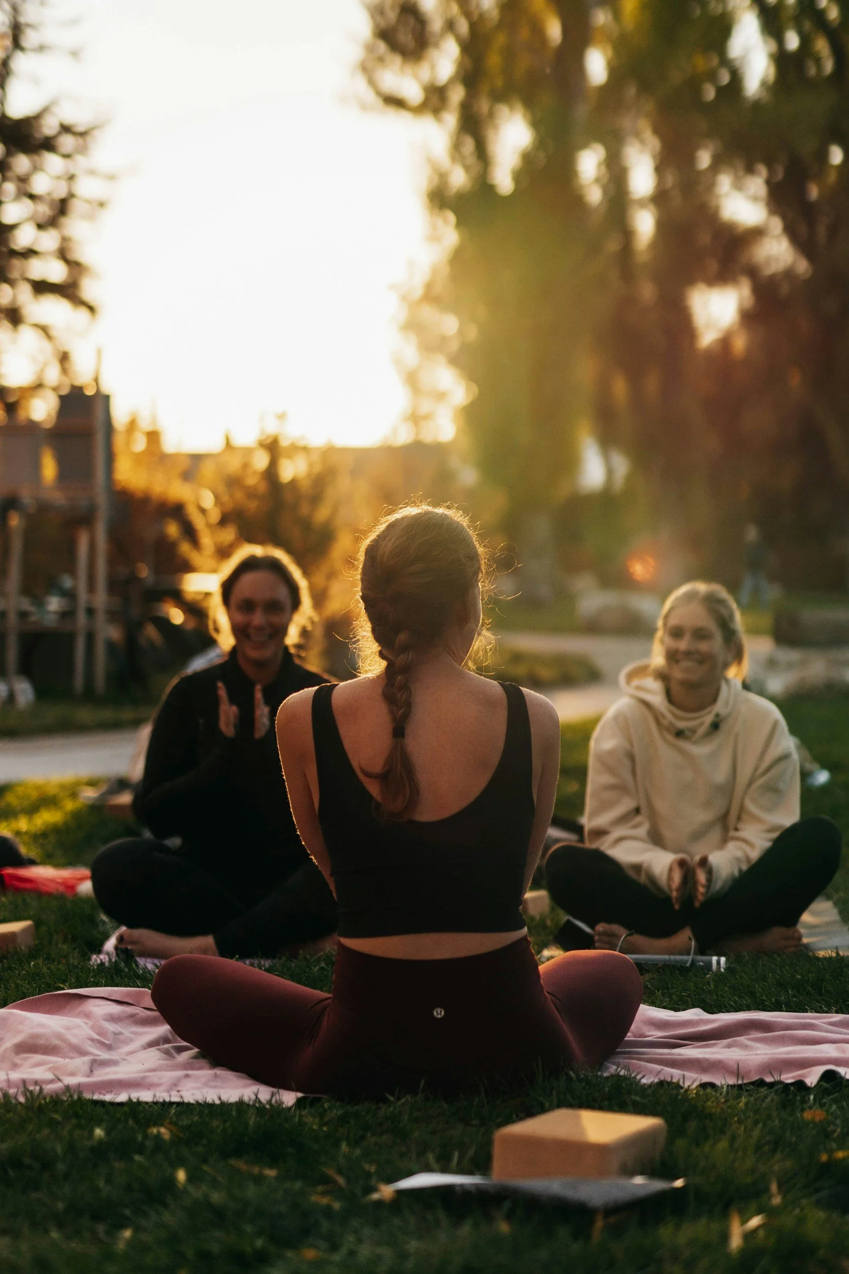 A group of people sitting on the grass in a circle outdoors during sunset, engaged in a meditation or yoga session. The focus is on a woman with a braid, sitting cross-legged on a pink blanket in the foreground, with other individuals smiling and participating in the activity.