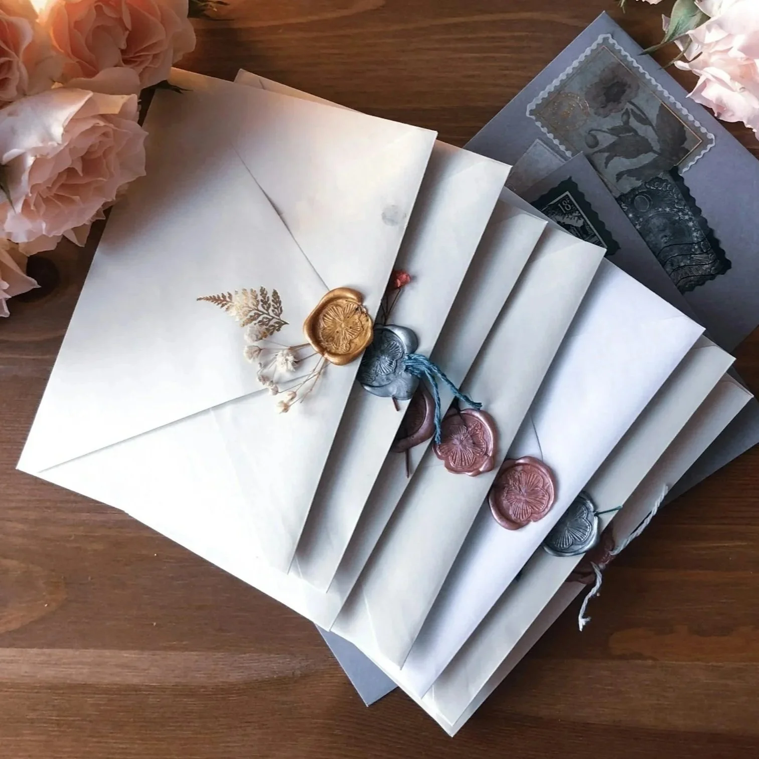 Several white envelopes with wax seals in gold, silver, and pink, arranged on a wooden surface with pale pink roses at the edges.