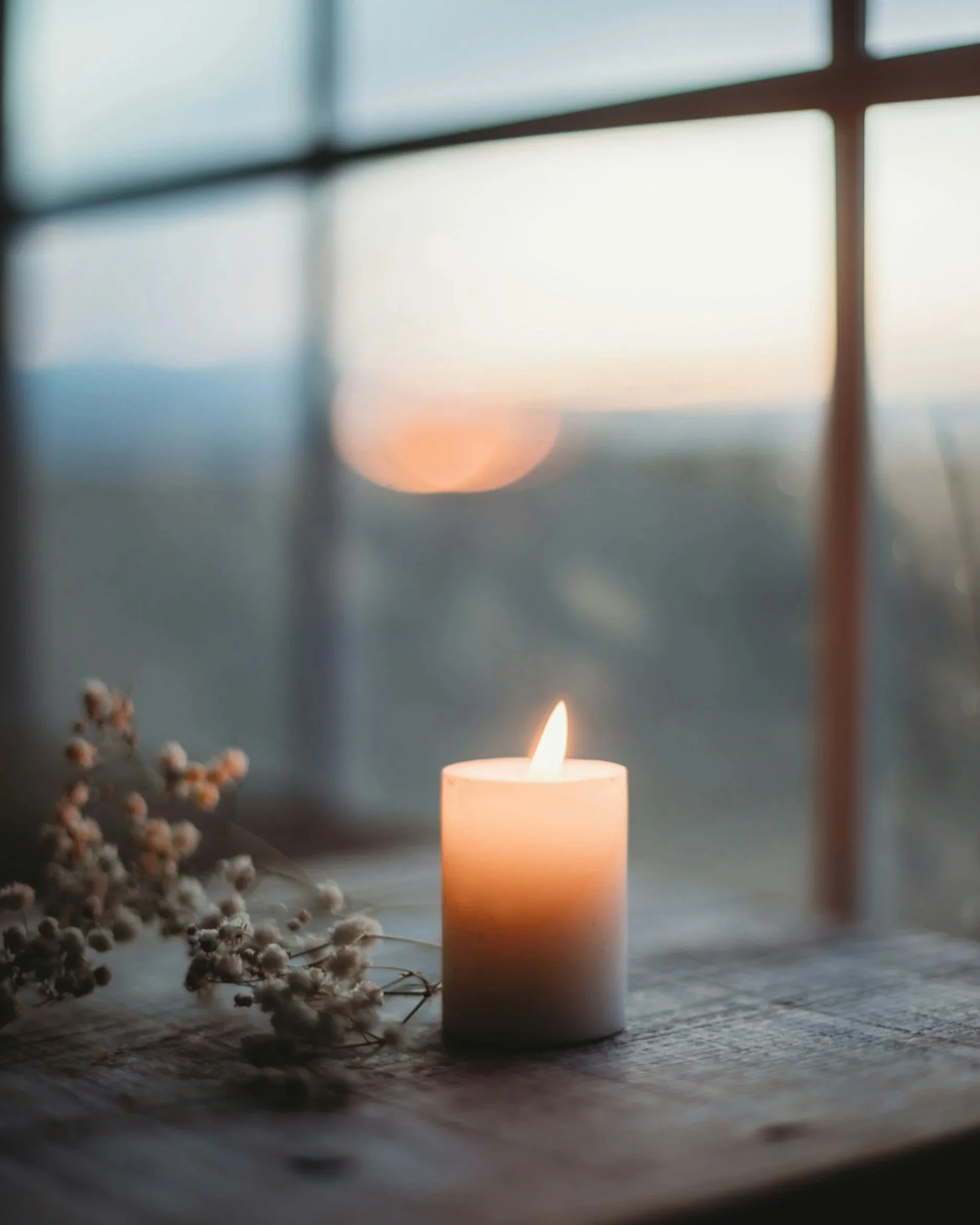 A lit white candle on a rustic wooden surface, with a blurred background of a window showing a sunset or sunrise sky, and a small bouquet of white flowers in the foreground.