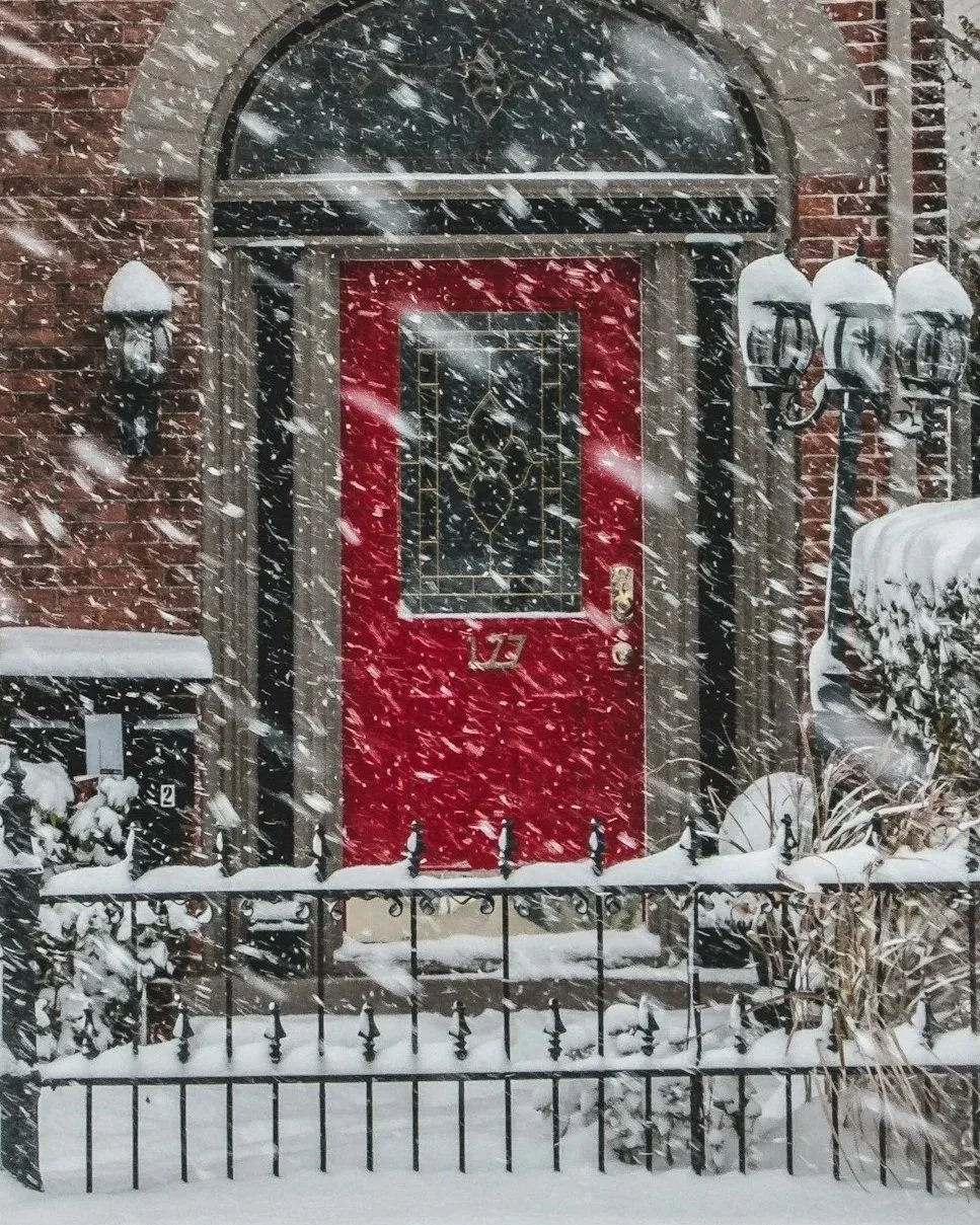 Snow falling in front of a brick house with a red door and decorative glass window, surrounded by snow-covered plants, lamp posts, and a black metal fence.