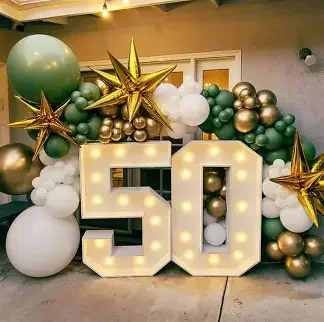 Decorative balloon arrangement with large illuminated number 50, gold, green, white, and silver balloons, topped with gold star balloons for a celebration.