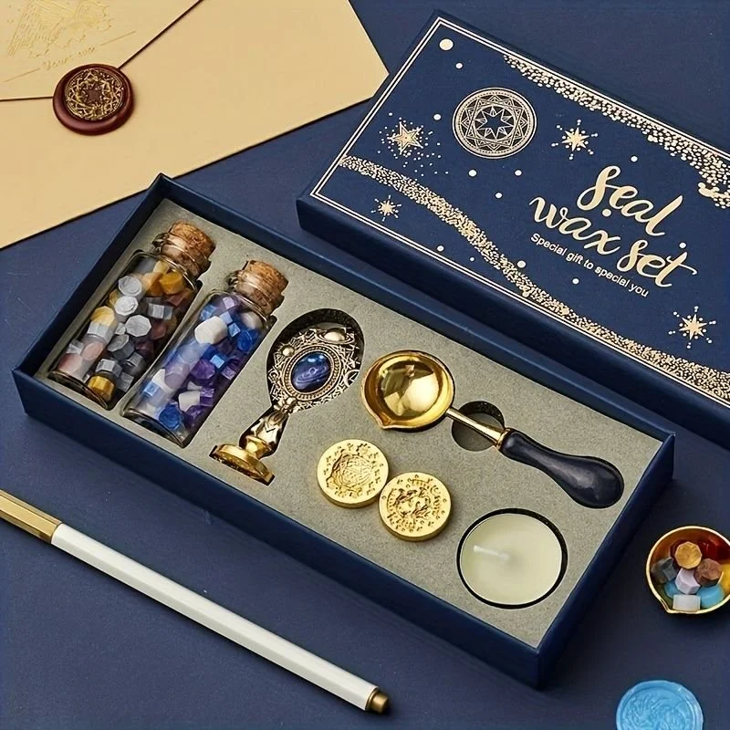 A gift set box labeled 'seal wax set' with gold and silver seals, metallic wax beads in small jars, a wax spoon, a sealing stamp, and a tea light candle for melting wax. The box and accessories are placed on a dark blue surface.