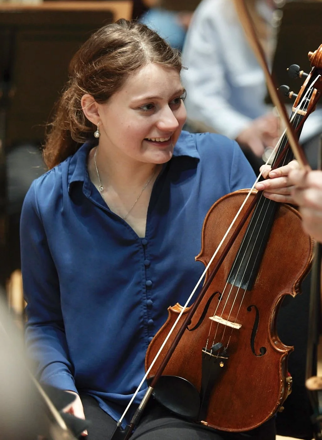 Young woman playing the violin during an orchestra rehearsal, wearing a blue blouse and earrings, smiling and looking at her instrument.
