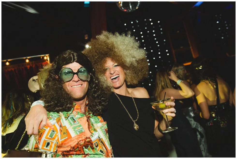 Rent retro camcorder for themed party Two people dressed in 70s disco attire at a party, smiling and enjoying drinks, with other partygoers in the background.