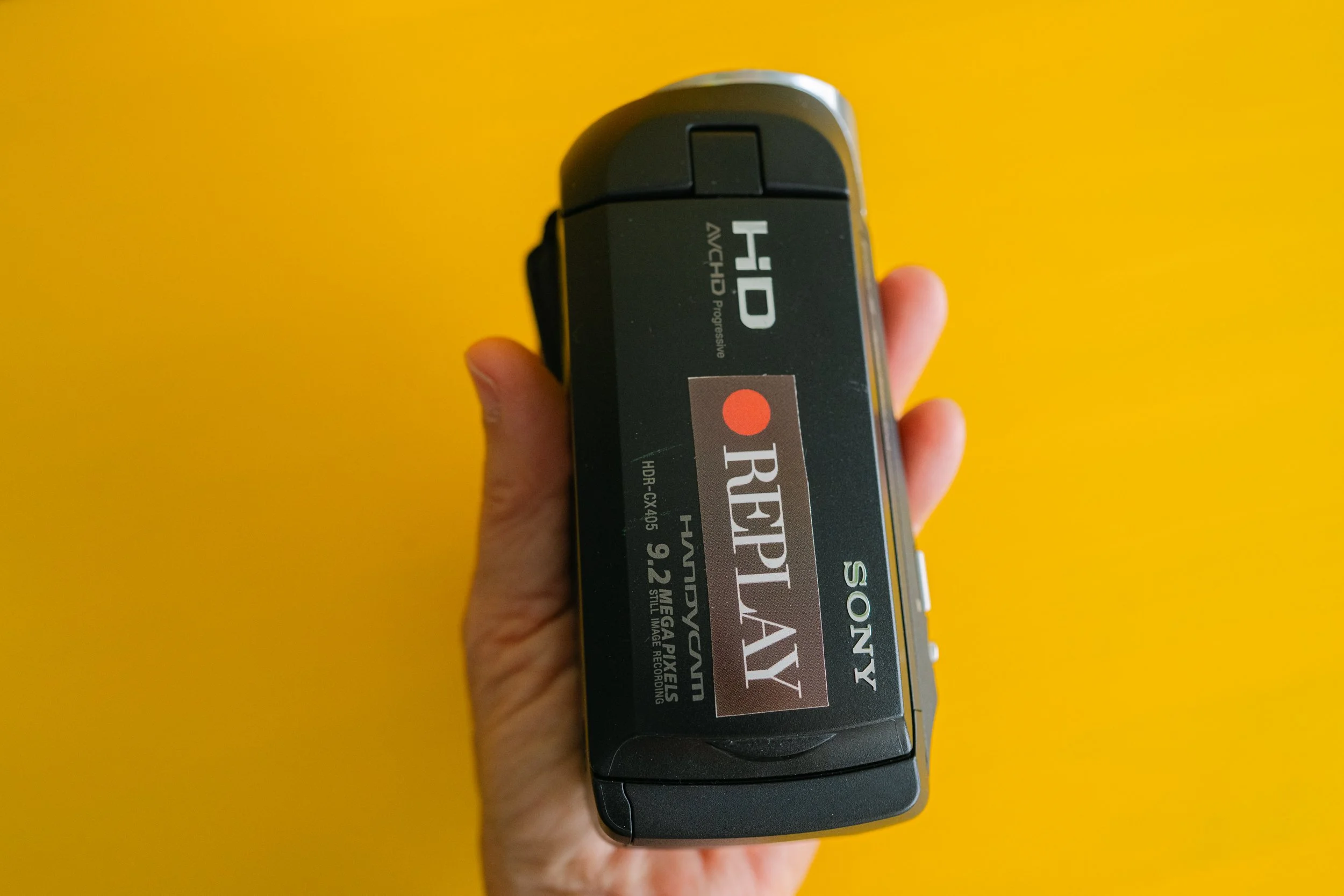 A hand holding a Sony HD camcorder with a black body and a label, against a bright yellow background.