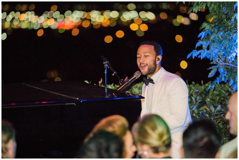 John Legend in a white tuxedo with a black bowtie singing at a piano during an outdoor night event, with string lights in the background and audience members in the foreground add retro camcorders to your parties