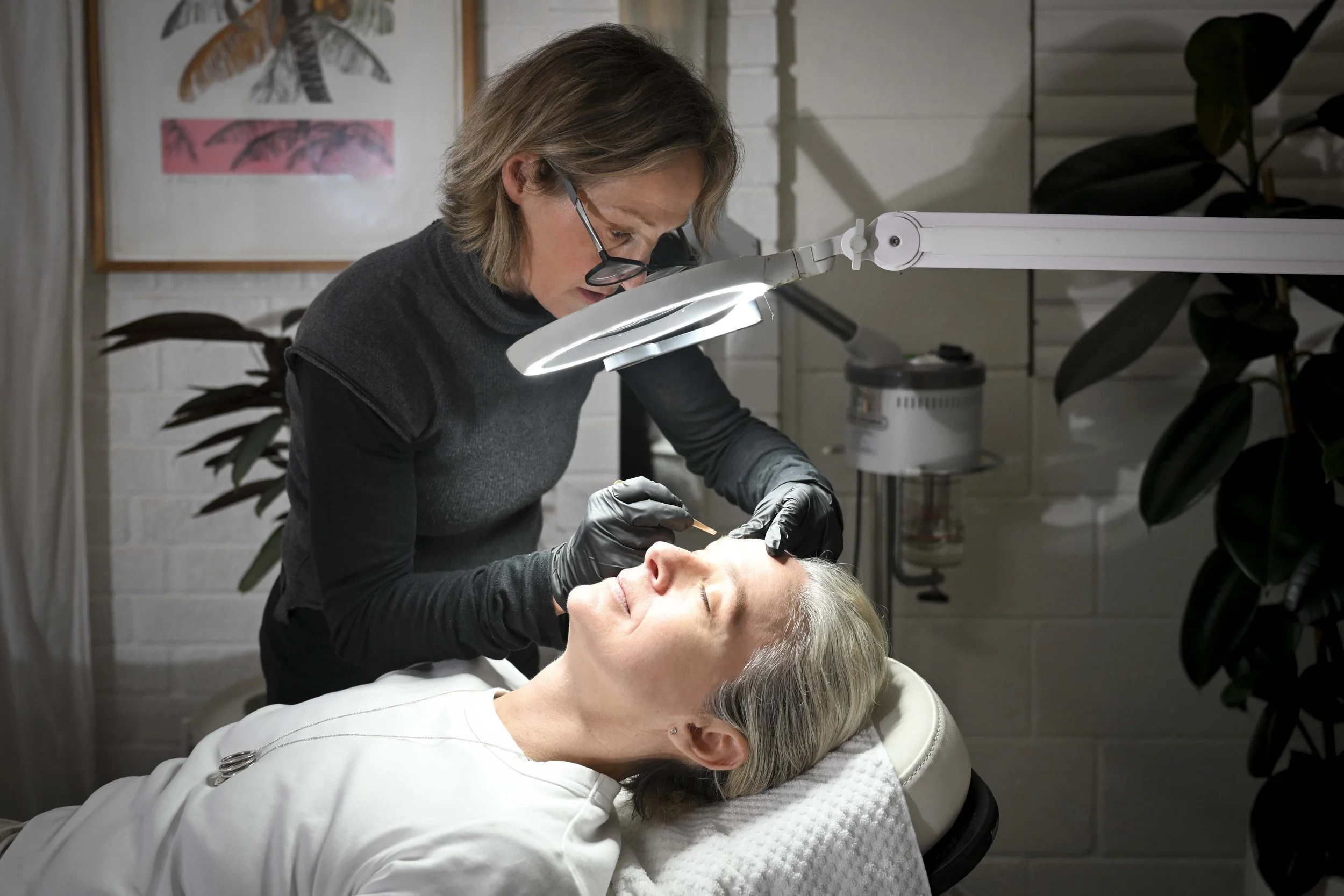 A cosmetologist performs a facial or skincare treatment on a woman with gray hair, who is lying down with her eyes closed, in a modern, well-lit beauty clinic or spa setting.