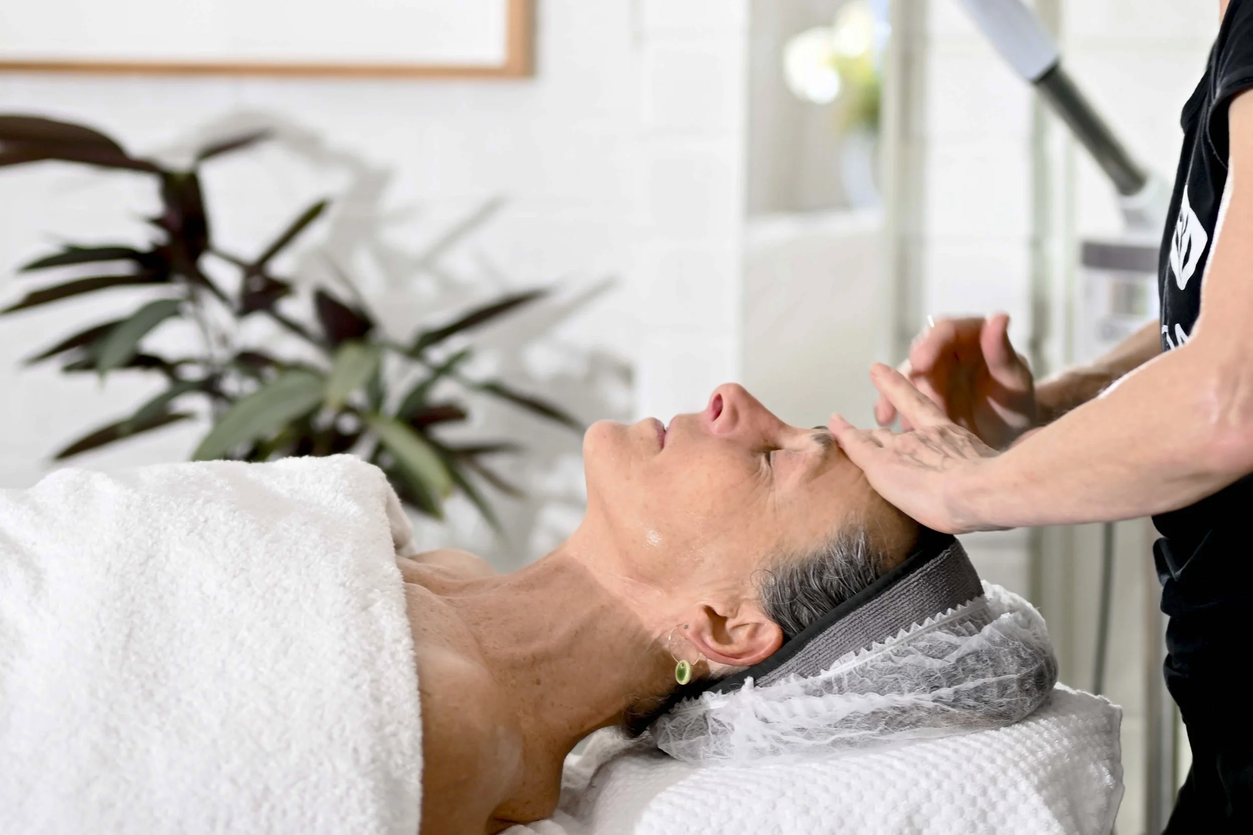 A woman lying on a spa bed receiving a facial treatment from a professional in a bright, modern spa or clinic room.