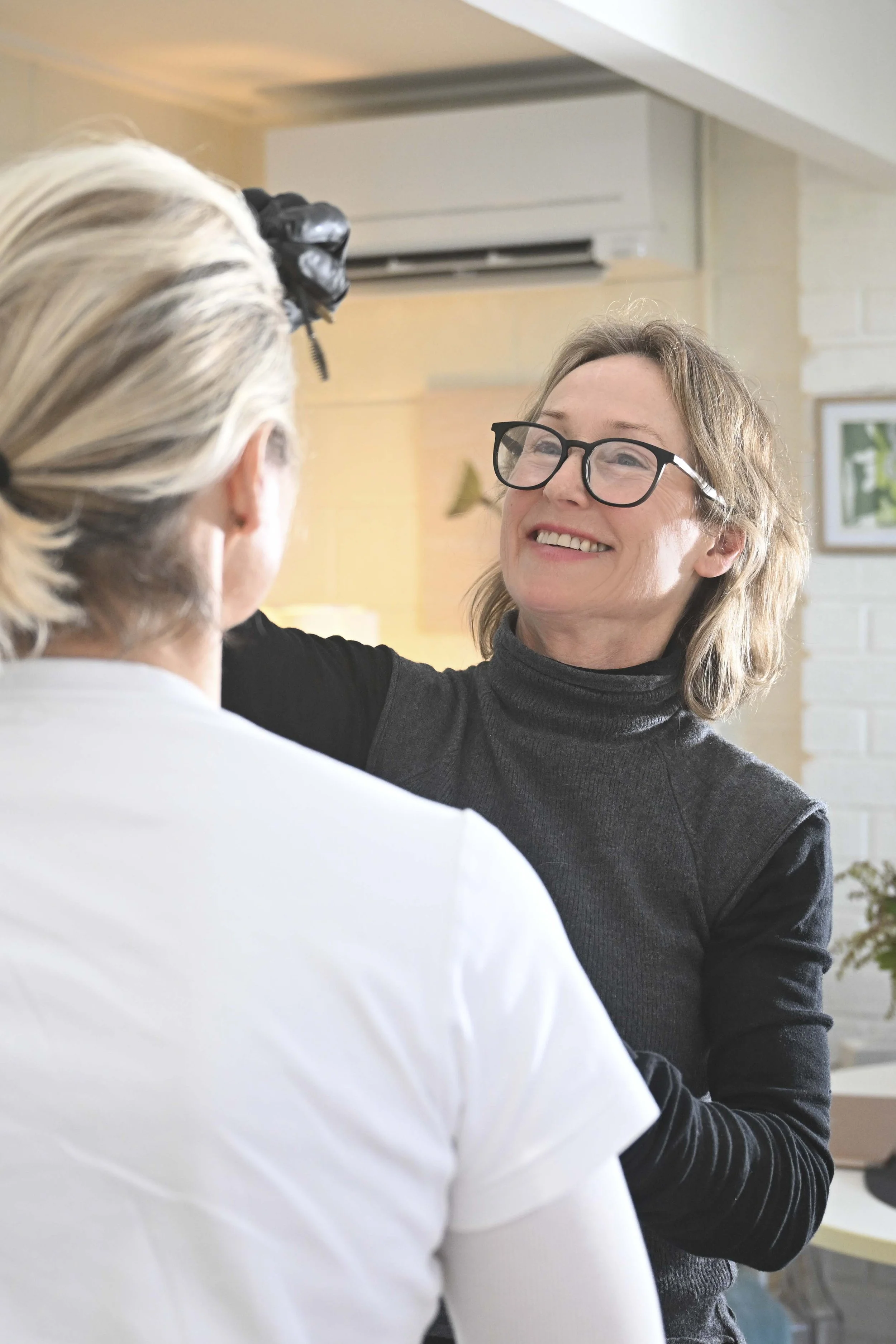 Two women, one with gray hair and glasses, smiling warmly and wearing a black turtleneck, are sharing a moment in a bright indoor space. The other woman, with blond hair in a bun and hair clip, is seen from behind.