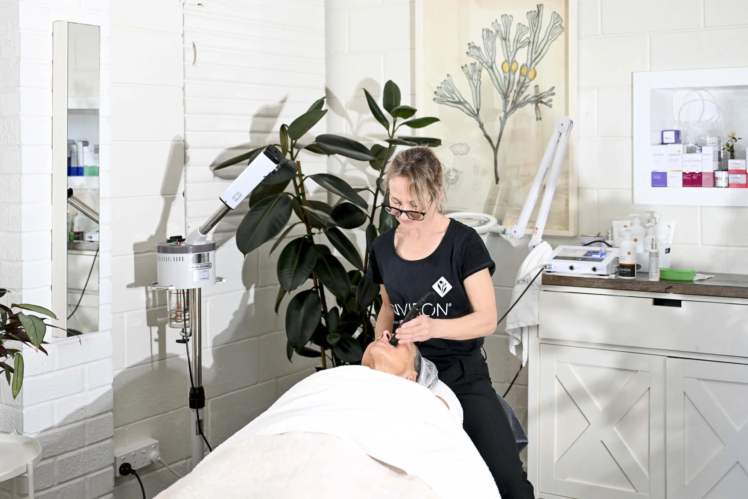 A skincare professional performing a facial treatment on a client in a modern spa or skincare clinic, with skincare products on shelves and a large leafy plant in the background.