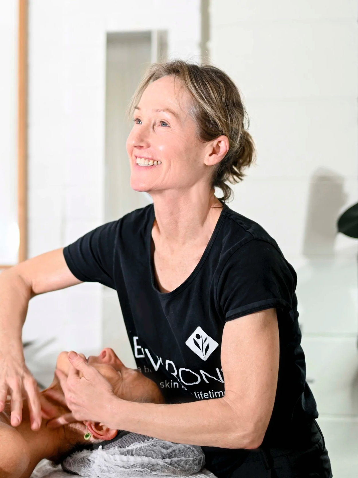 A smiling woman with short, light brown hair, wearing a black t-shirt, giving a facial massage or skincare treatment to a person lying down. The setting appears to be a skincare or wellness clinic.