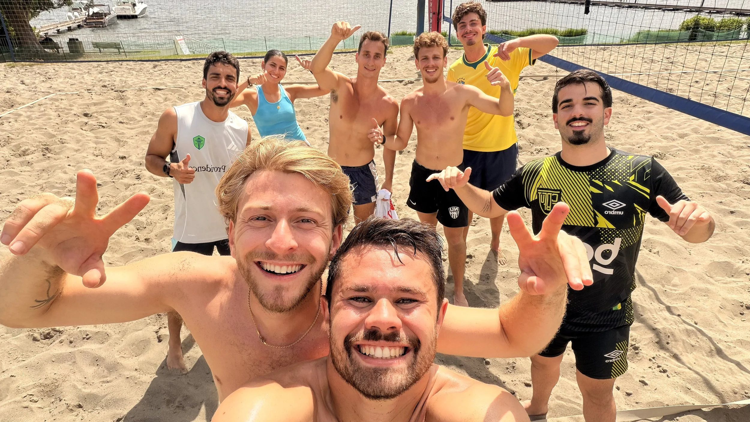 A group of eight friends taking a selfie on a sandy beach volleyball court, smiling and celebrating.
